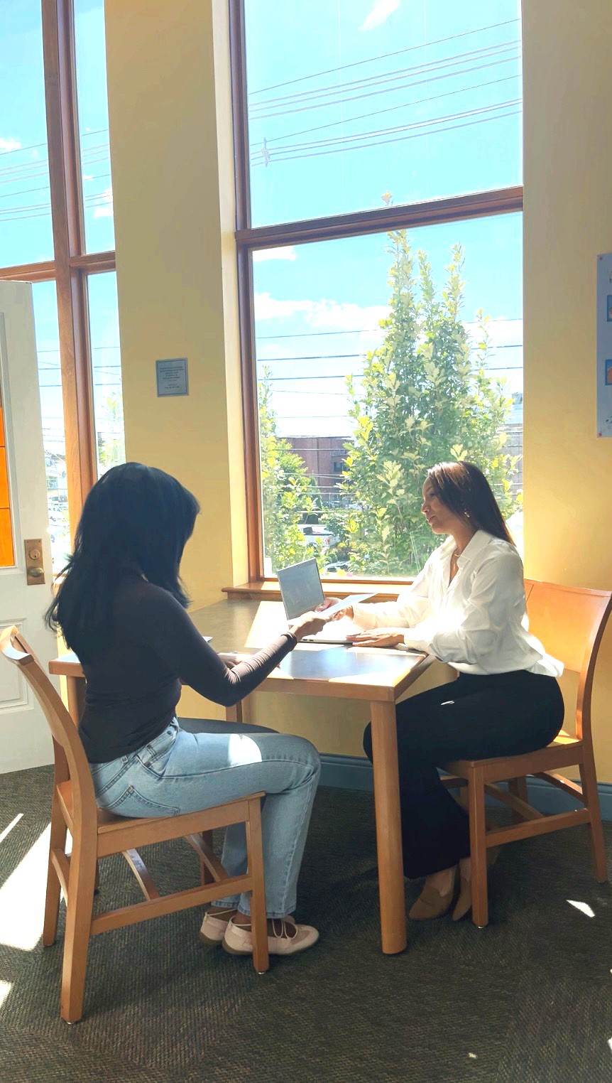 Two women sit at a table near large windows, working on laptops with trees and sky outside.