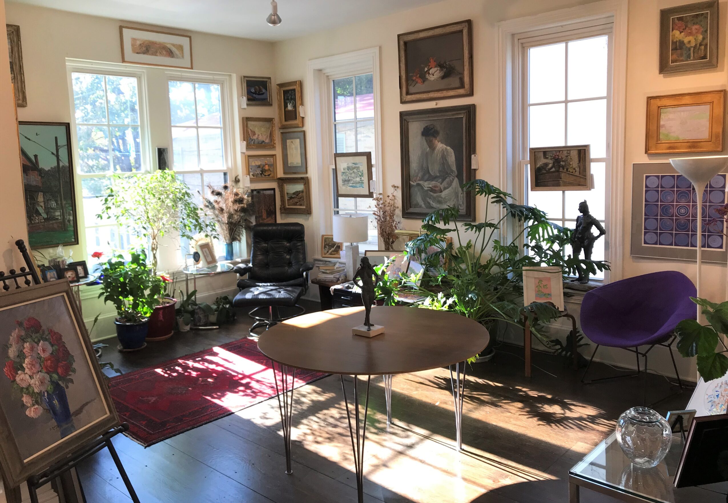 Sunlit room with framed artwork, plants, a table, and chairs, decorated with a red rug and various paintings on walls.