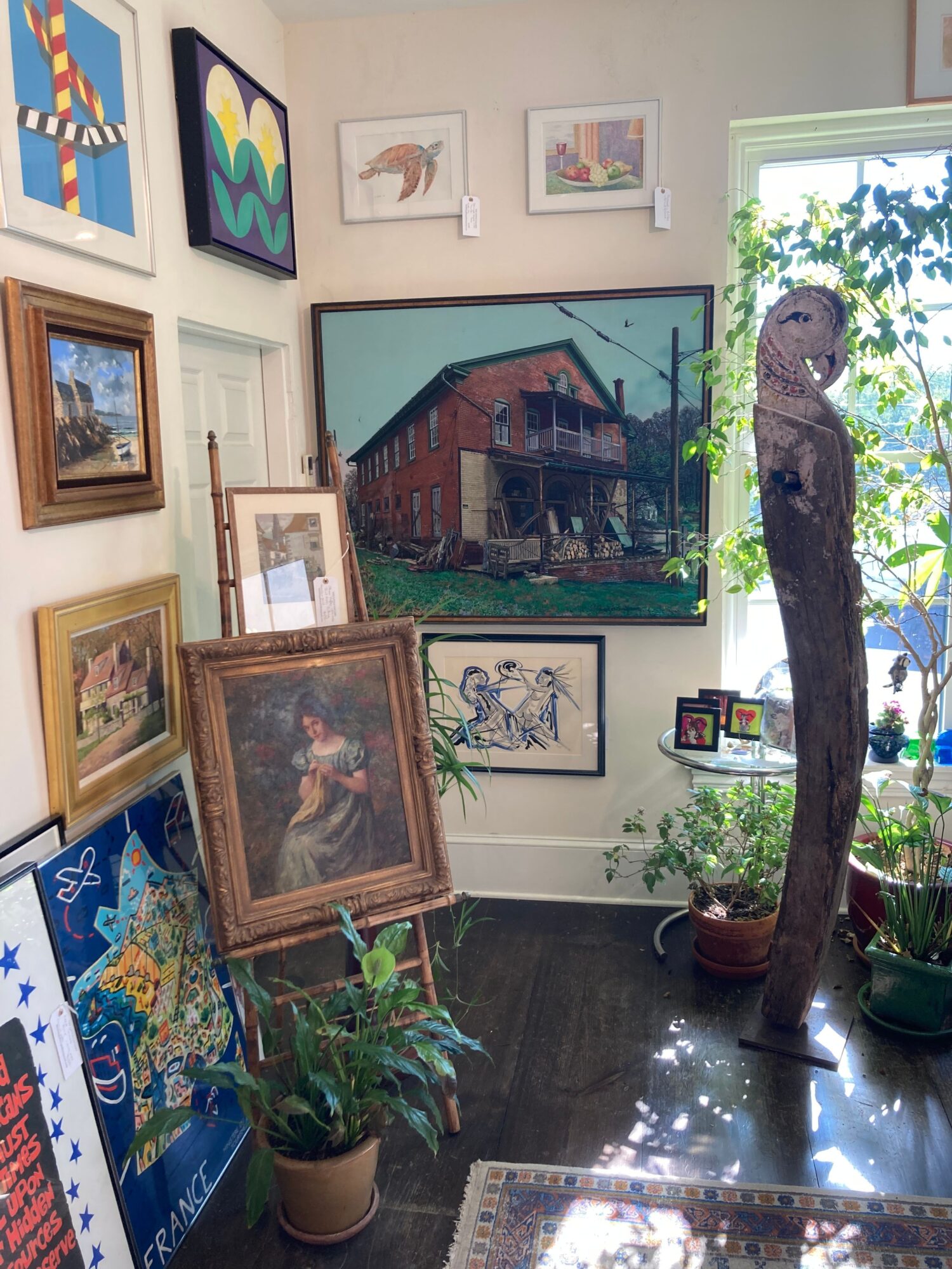 Room with framed artwork, potted plants, and a window with sunlight, viewed from the side. Several paintings and photographs are displayed on the wall and on surfaces.