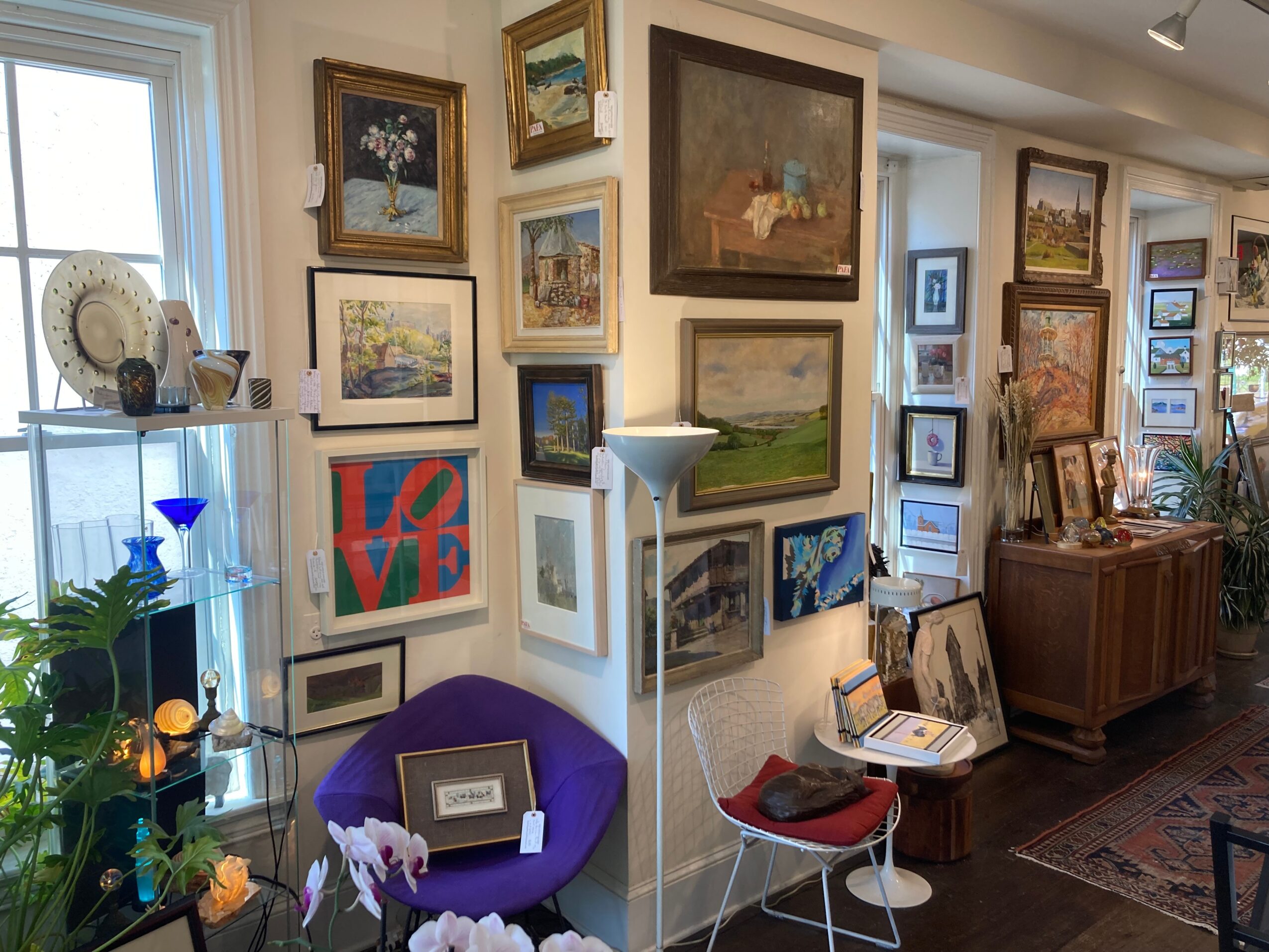 Gallery wall with framed paintings and photographs in a room with a purple chair, floor lamp, and wooden cabinet.