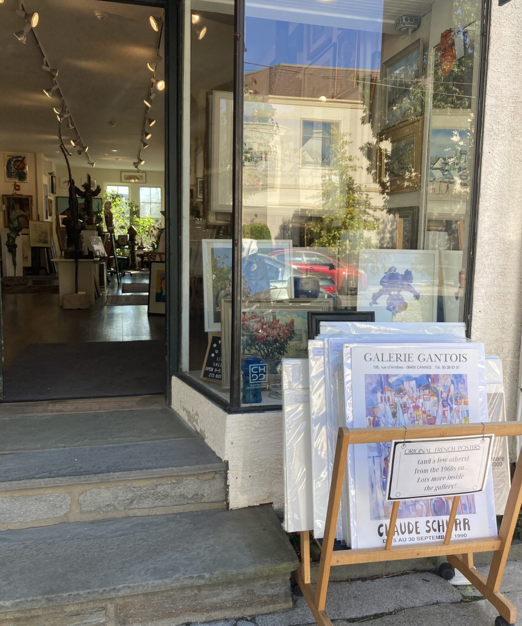 View of an art gallery entrance with glass door, display of paintings outside, and a signboard with artwork inside.