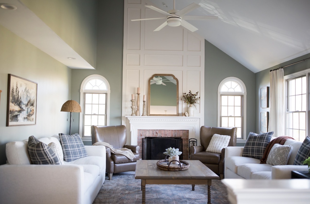 Living room with sofas, armchair, fireplace, mirror, and windows, decorated in neutral tones with plants and artwork.