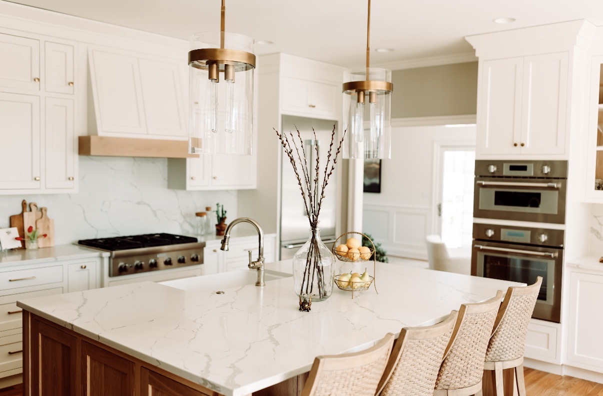 Bright kitchen with white cabinets, marble island, and pendant lights, featuring a stove, oven, and bar stools.