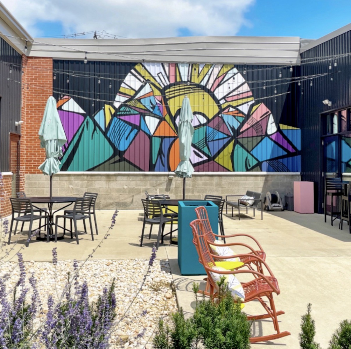 Outdoor patio with tables, chairs, and a colorful mountain and sun mural on the wall, under a partly cloudy sky.