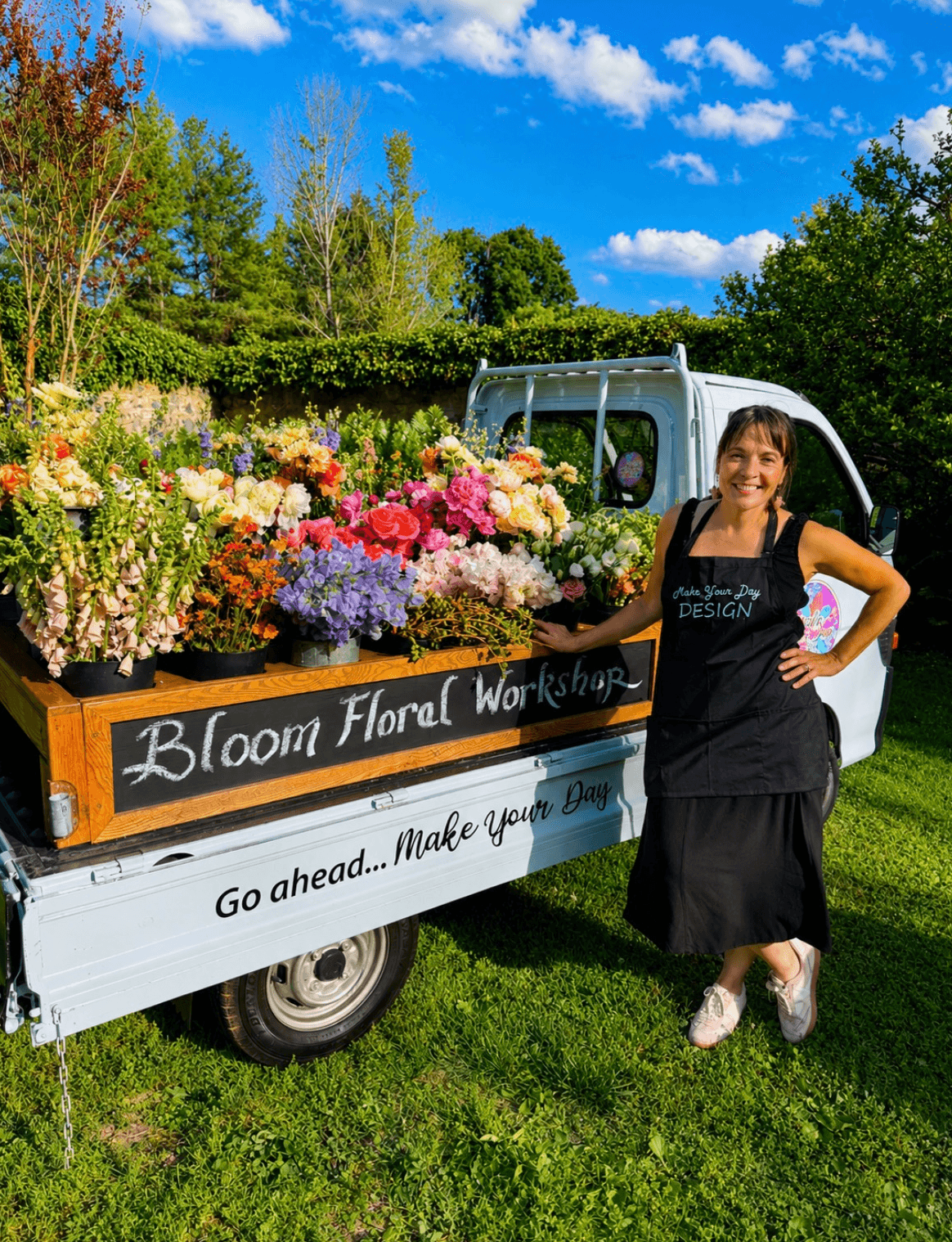 Woman in black apron standing next to a flower-filled truck with a sign for Bloom Floral Workshop outdoors on a sunny day.