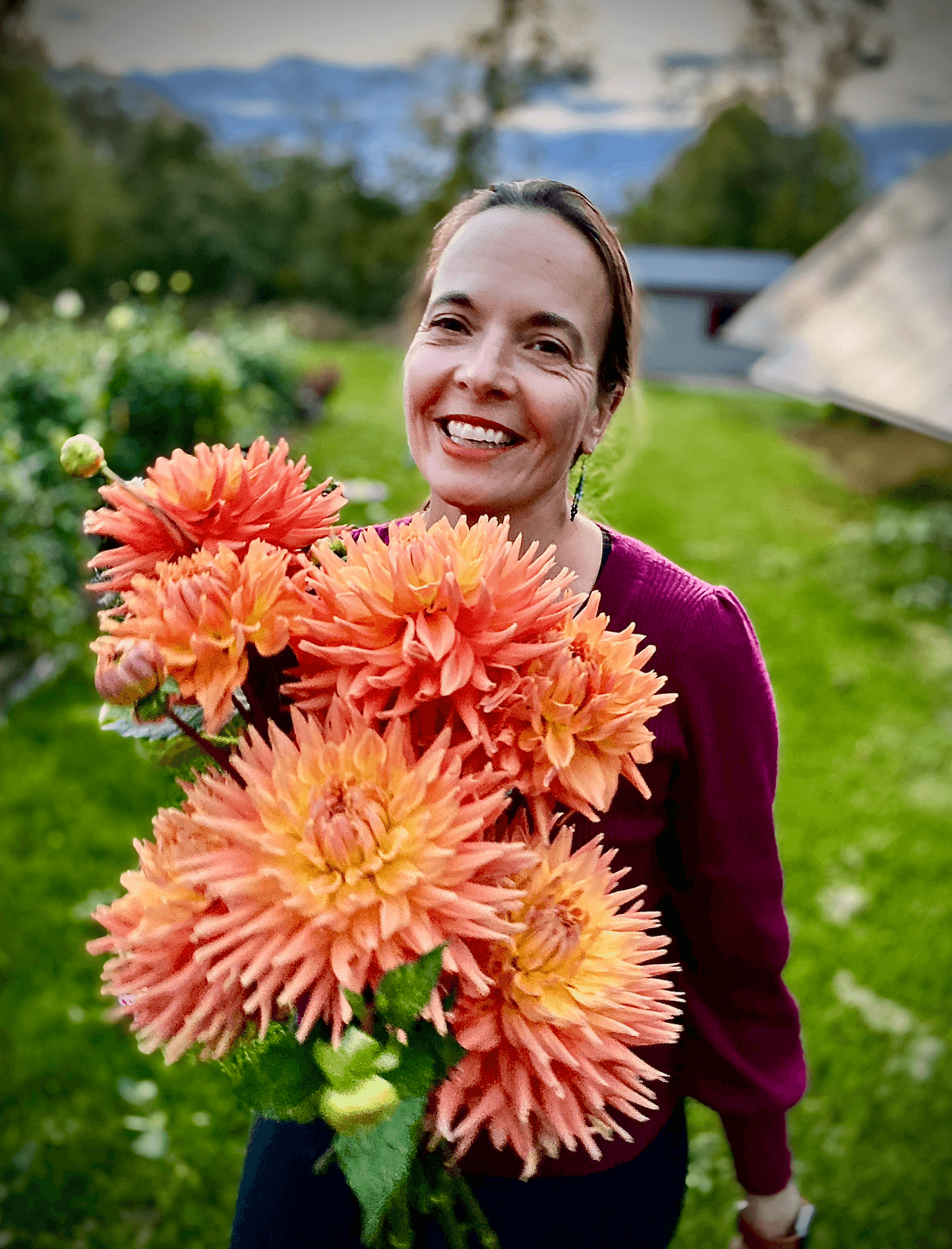Smiling woman holding large orange and yellow flowers outdoors with greenery and a building in the background.
