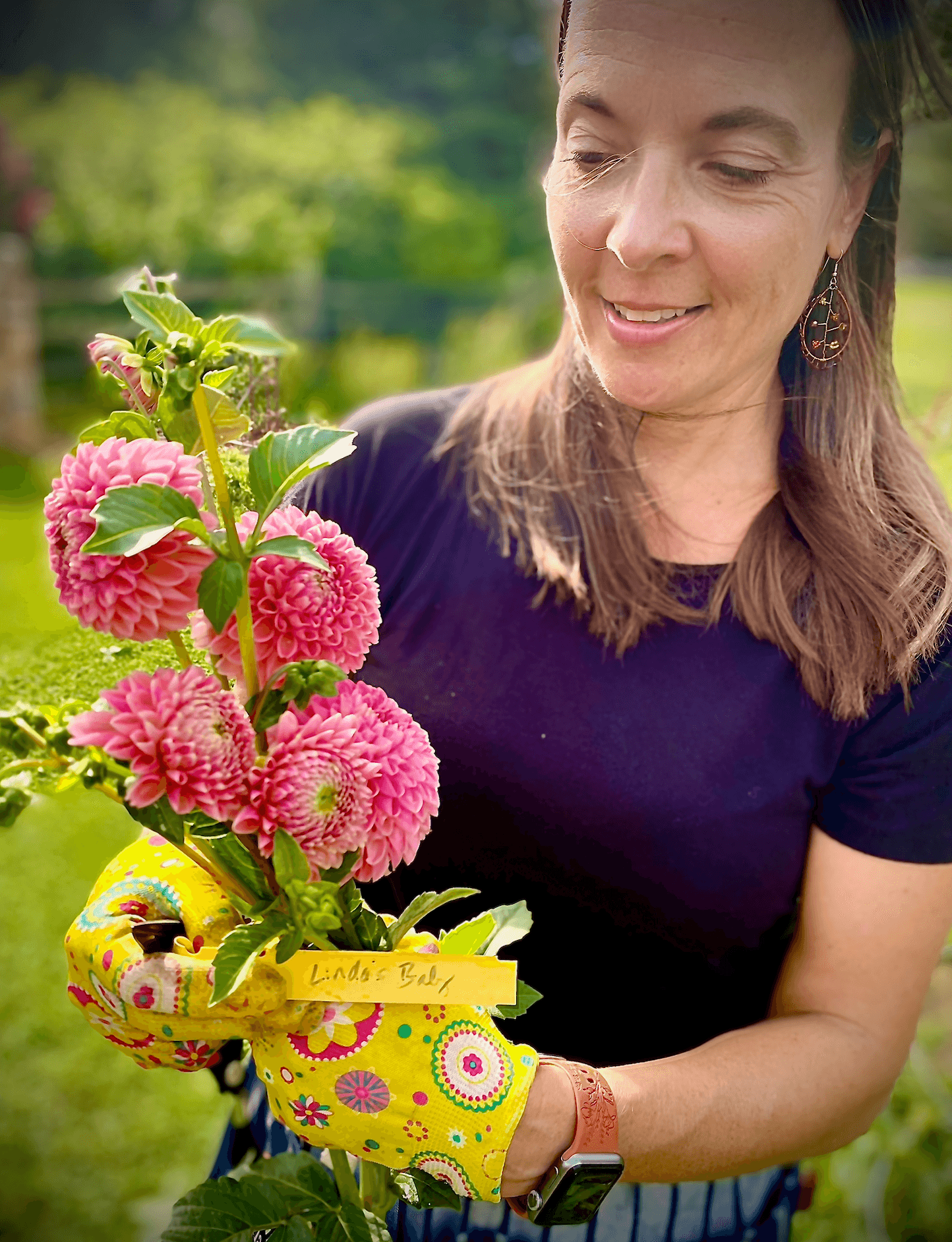 Woman holding a bouquet of pink flowers outdoors, wearing a dark shirt and yellow gardening glove, smiling.