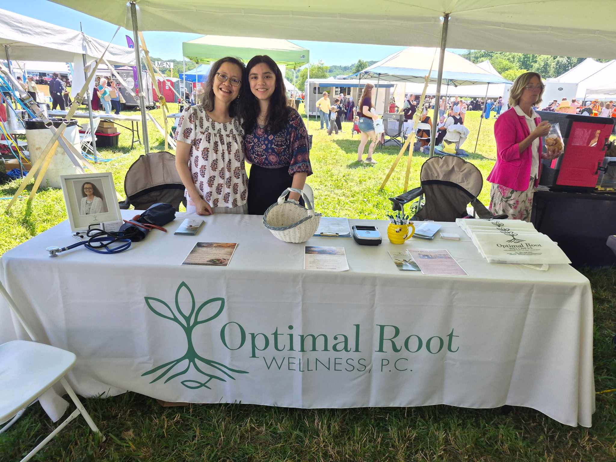 Two women stand behind a table with a banner reading 'Optimal Root Wellness P.C.' at an outdoor event, tents and people in background.