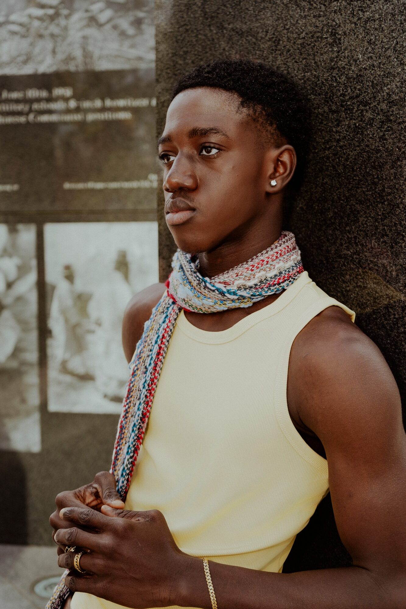 Young person with short hair, wearing a sleeveless top and colorful scarf, standing outdoors near a tree.