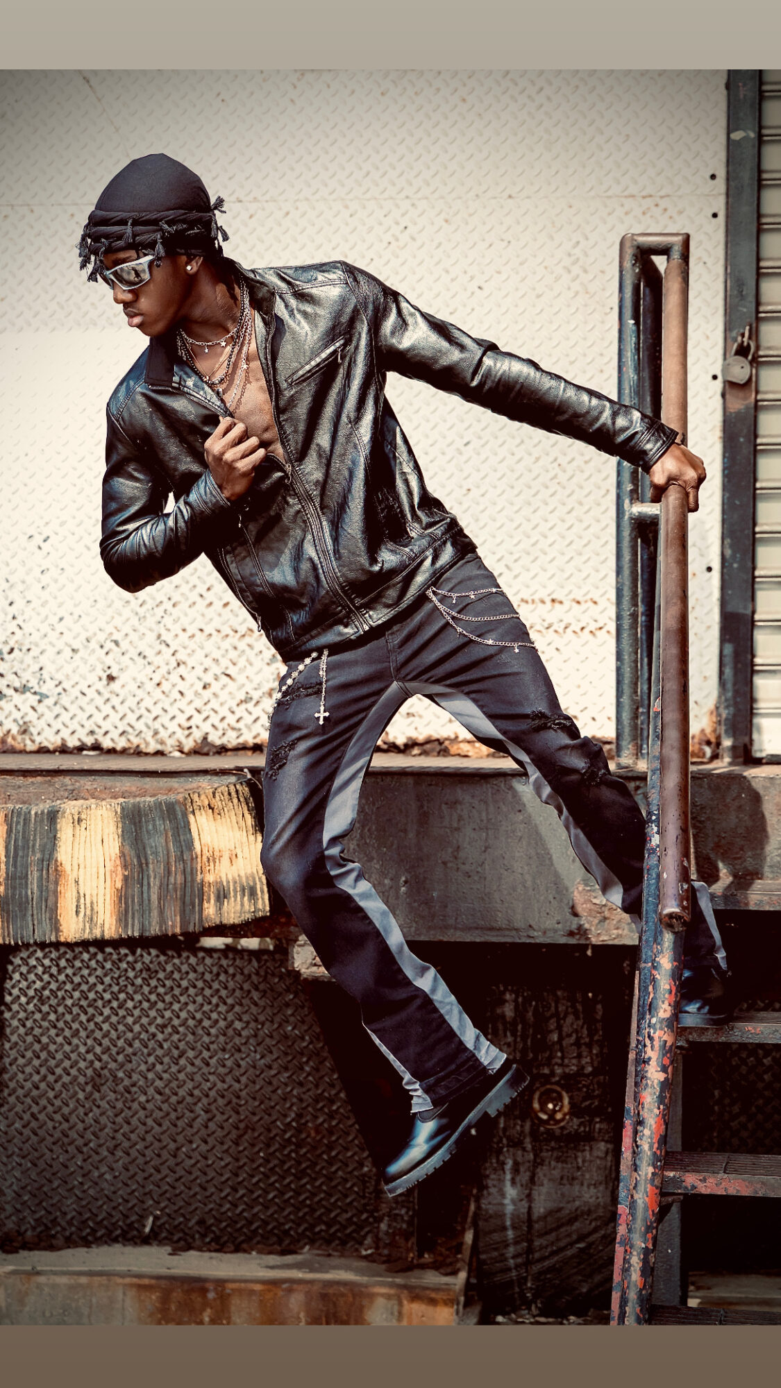 Person in black leather jacket and pants climbing a metal ladder outdoors, wearing glasses and a cap.