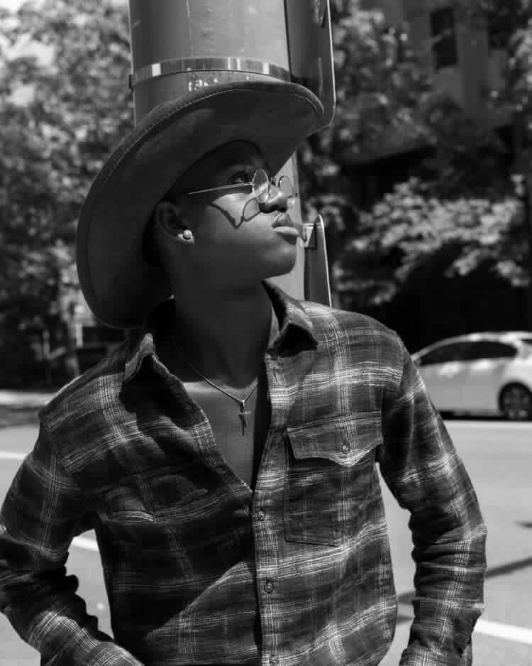 Person wearing a hat and glasses, looking upward outdoors, with trees and cars in background.
