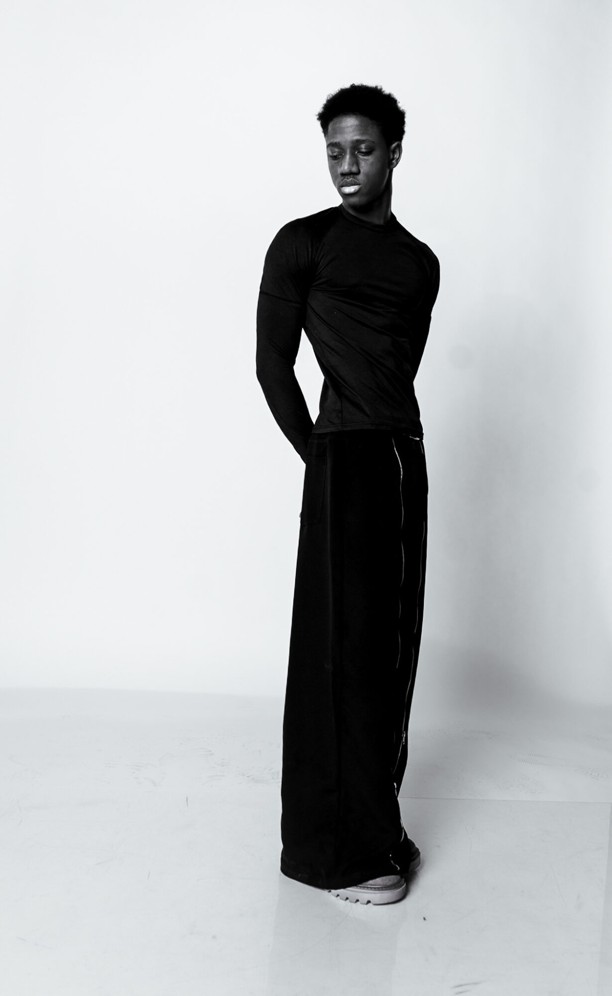 A person with short hair wearing a long-sleeved black top and wide black pants, standing against a plain background.