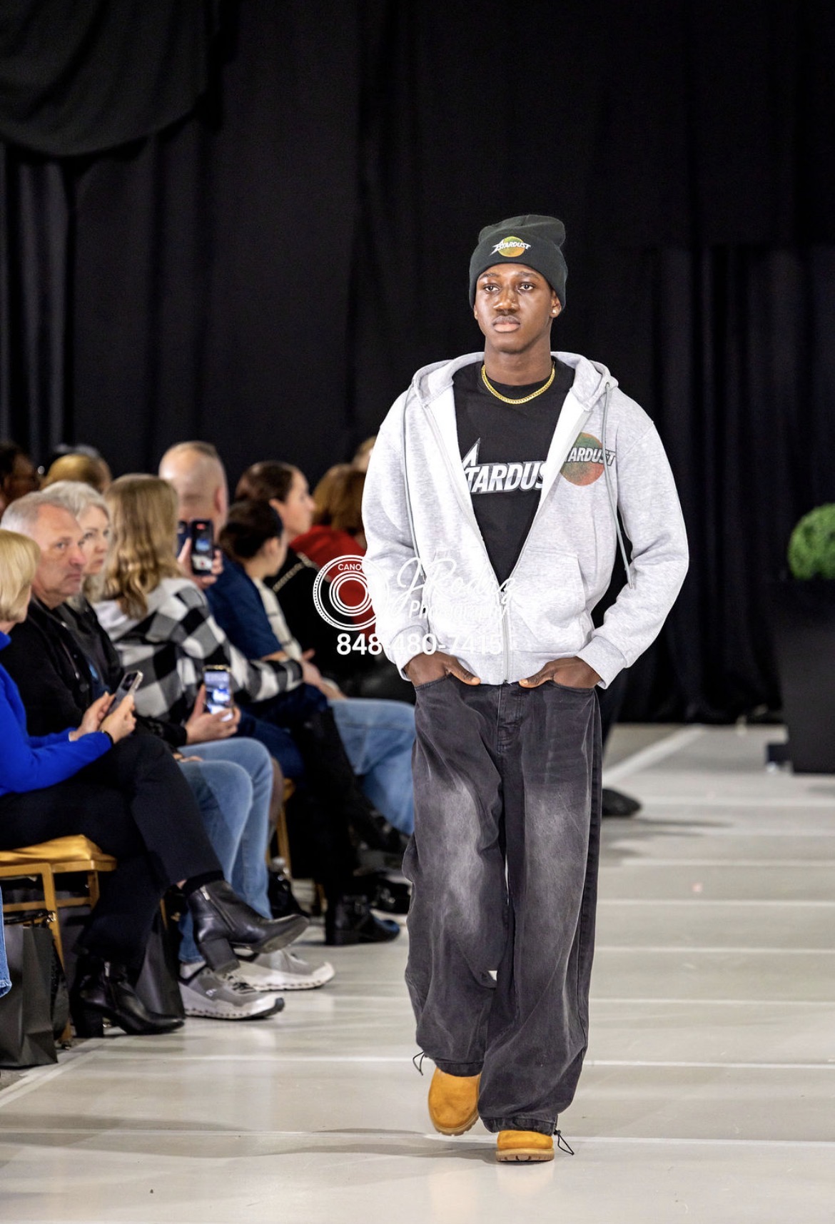 Young male model walking on runway in gray hoodie, black pants, yellow shoes, black beanie, audience seated on sides.