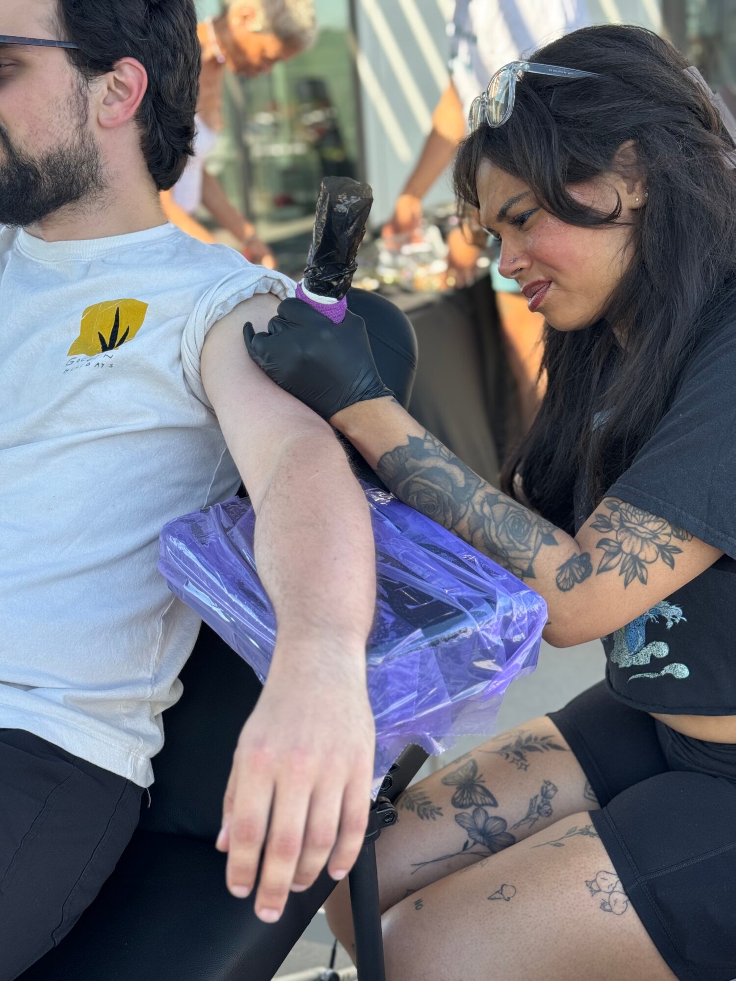 Woman with tattoos receives a tattoo on man's arm at an outdoor event, both appear focused.