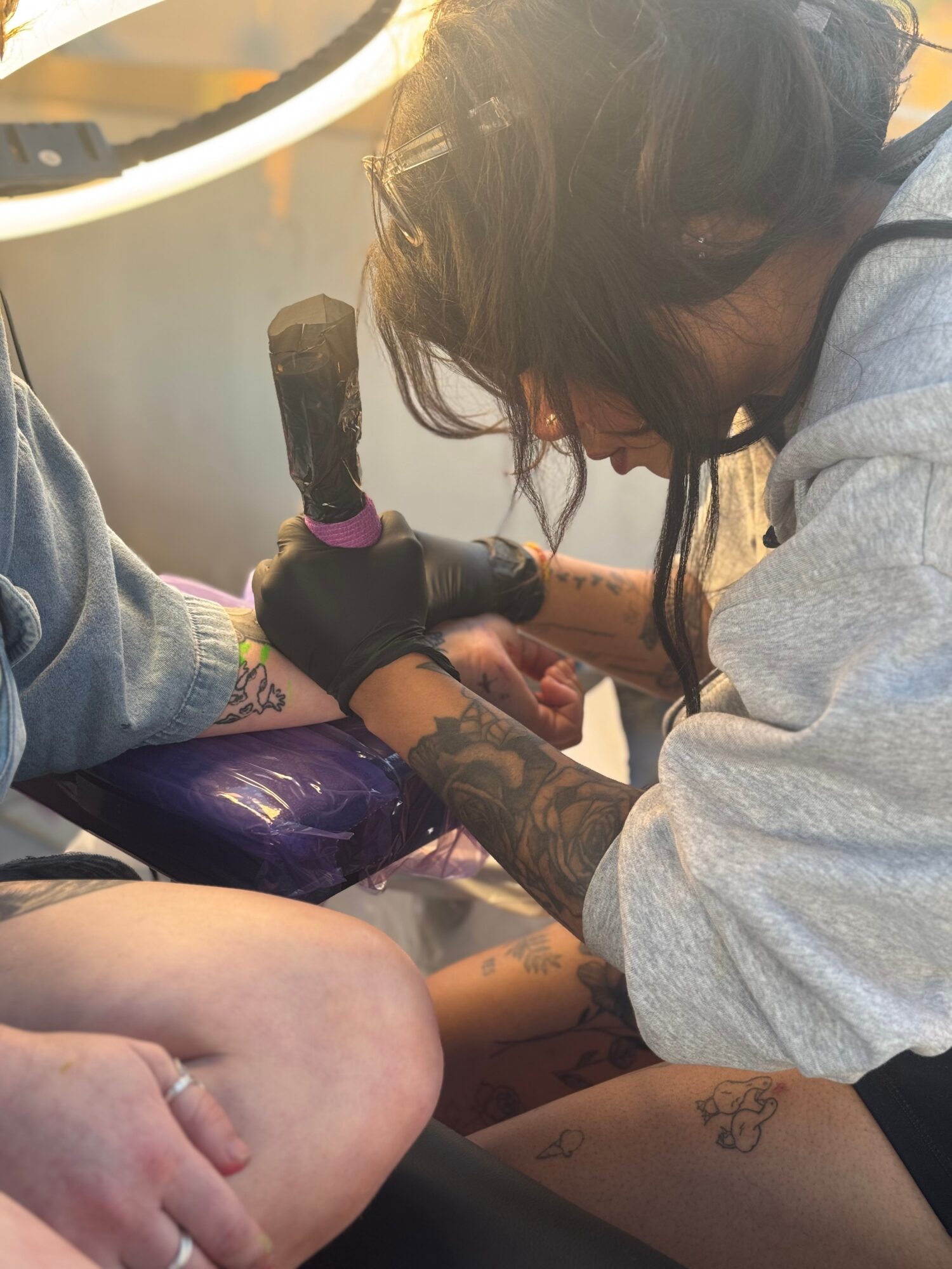 Person with tattoos and black gloves tattooing another person's leg with a tattoo machine, in a close-up view.