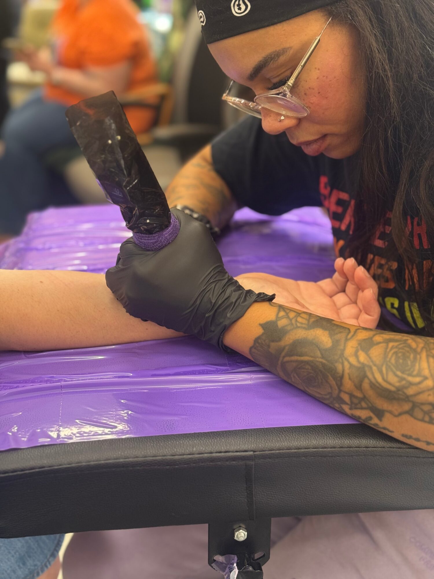 Tattoo artist with glasses and headband tattooing a person's arm, using a tattoo machine, on a purple surface.