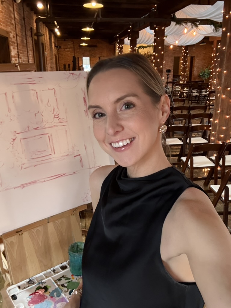 Smiling woman in black sleeveless dress taking a selfie in a decorated indoor venue with tables and string lights.