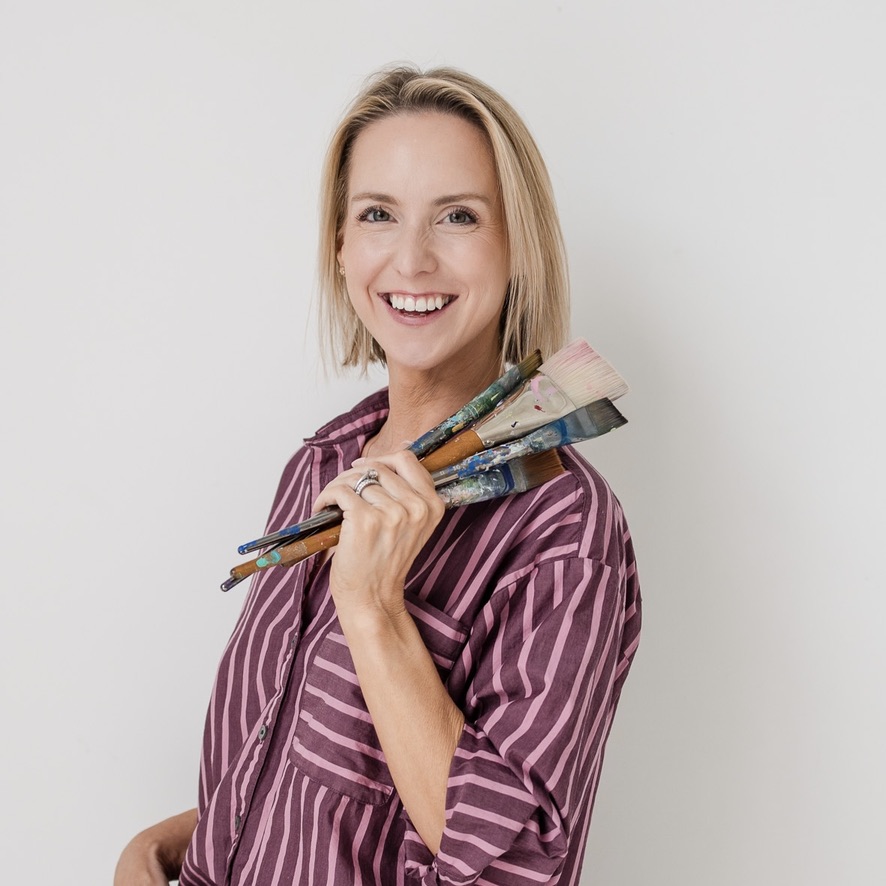 Smiling woman with shoulder-length blonde hair holding paintbrushes, wearing a striped purple shirt, standing against a plain white background.