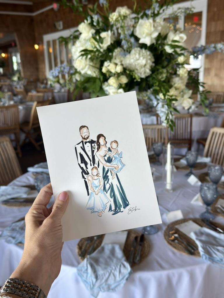 Hand holding a wedding illustration card in front of a decorated table with a large floral centerpiece, in a banquet hall.