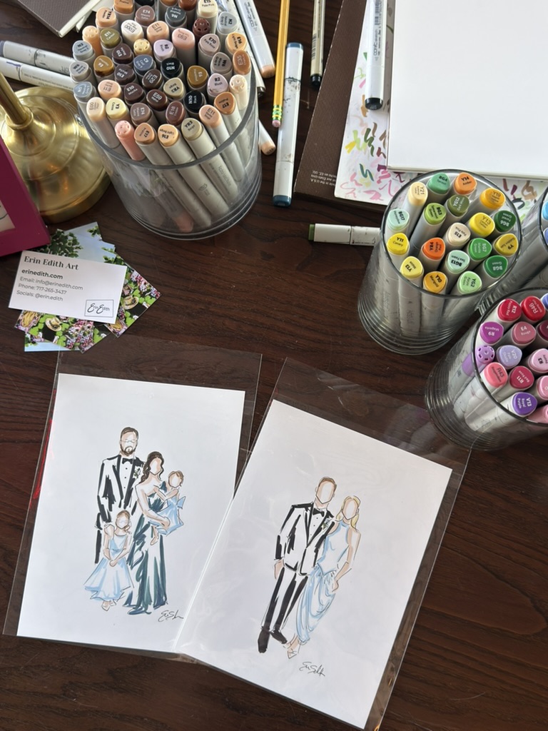 Two fashion sketches on paper, surrounded by colorful markers and pens on a wooden table.