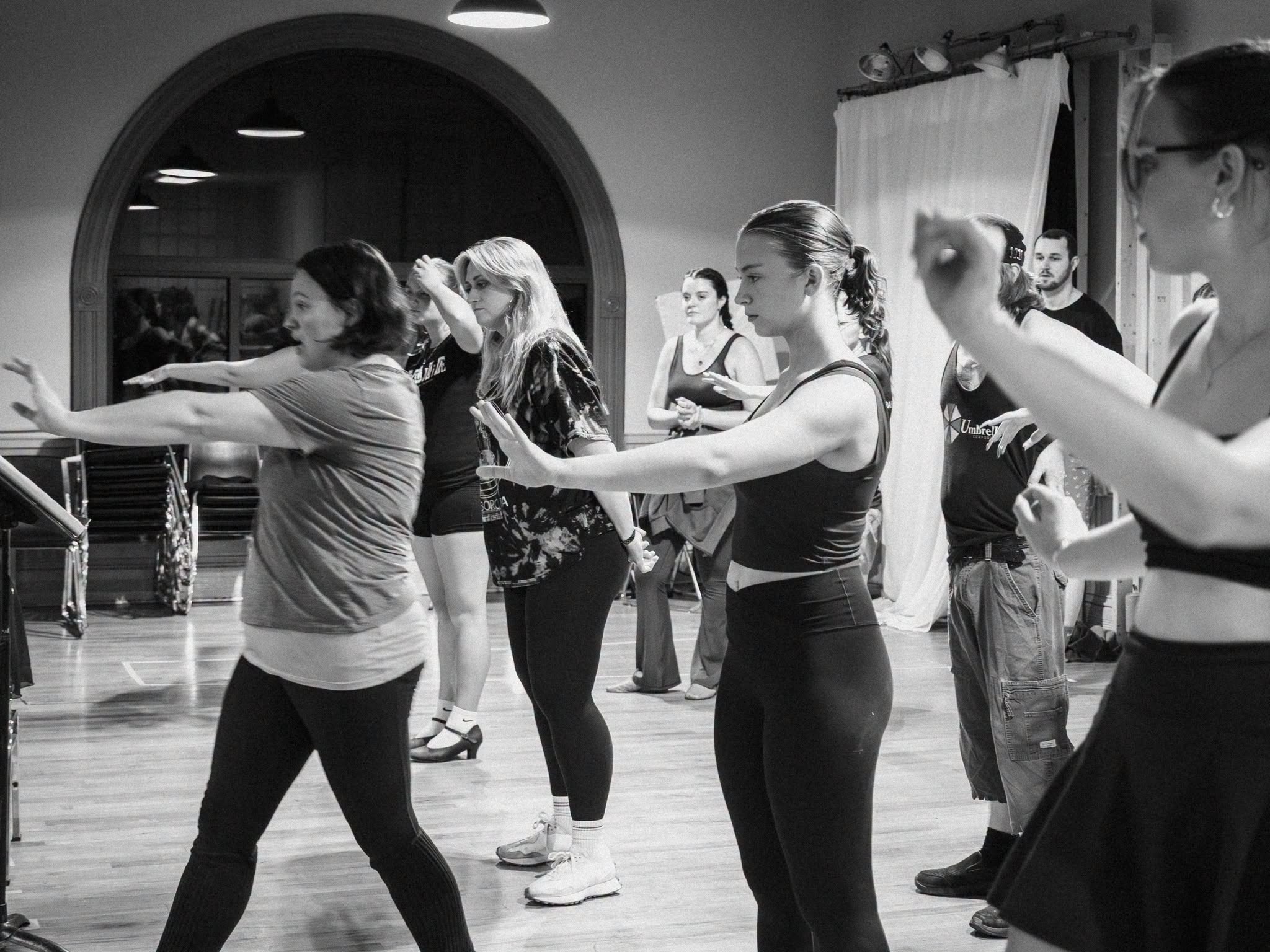 Group of women practicing dance moves in a studio, standing in a line with arms extended forward, focused on their movements.