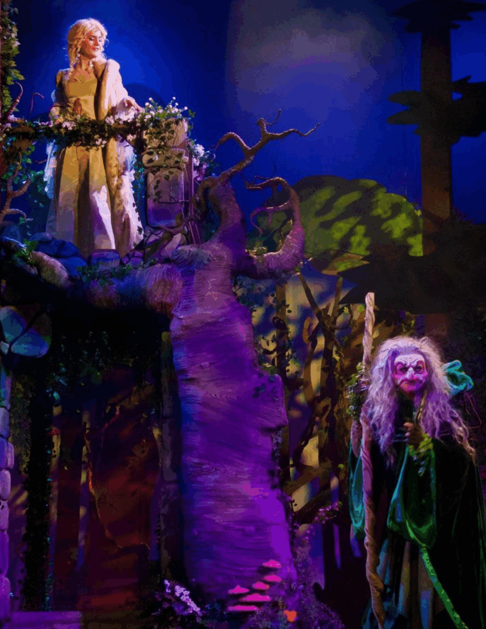 Stage scene with two characters, one on a tree branch and another in front, surrounded by colorful scenery and lighting.
