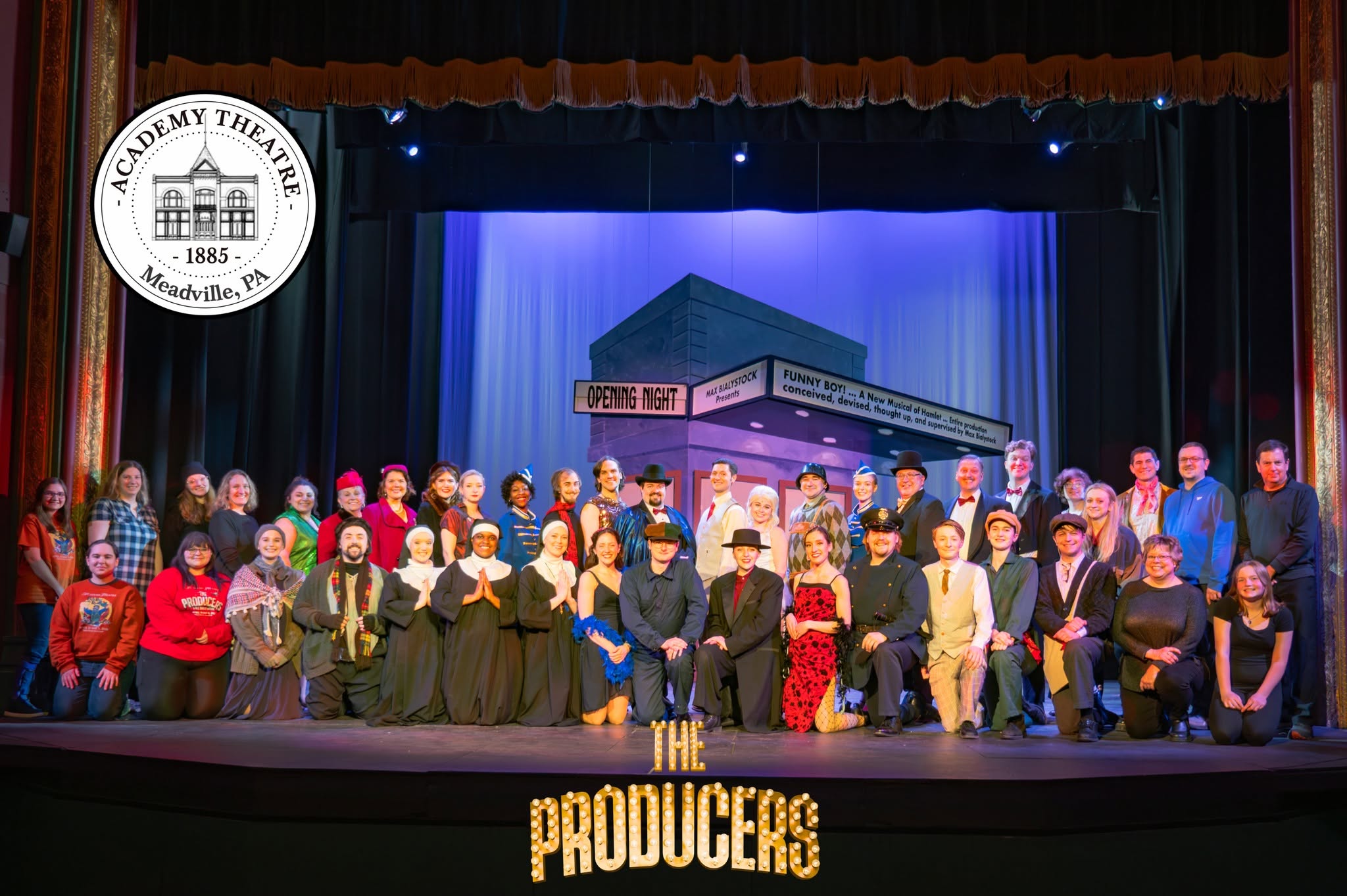 Group of people on stage in front of a theater set, with a sign reading 'The Producers' and a theater logo in the top left corner.