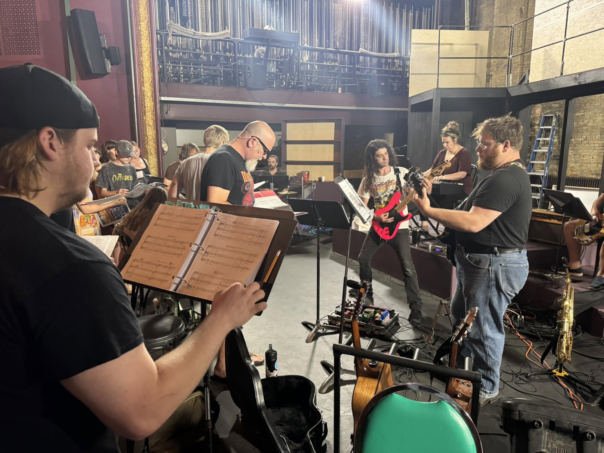 Group of musicians rehearsing in a studio, with a person reading sheet music in the foreground and others playing instruments in the background.
