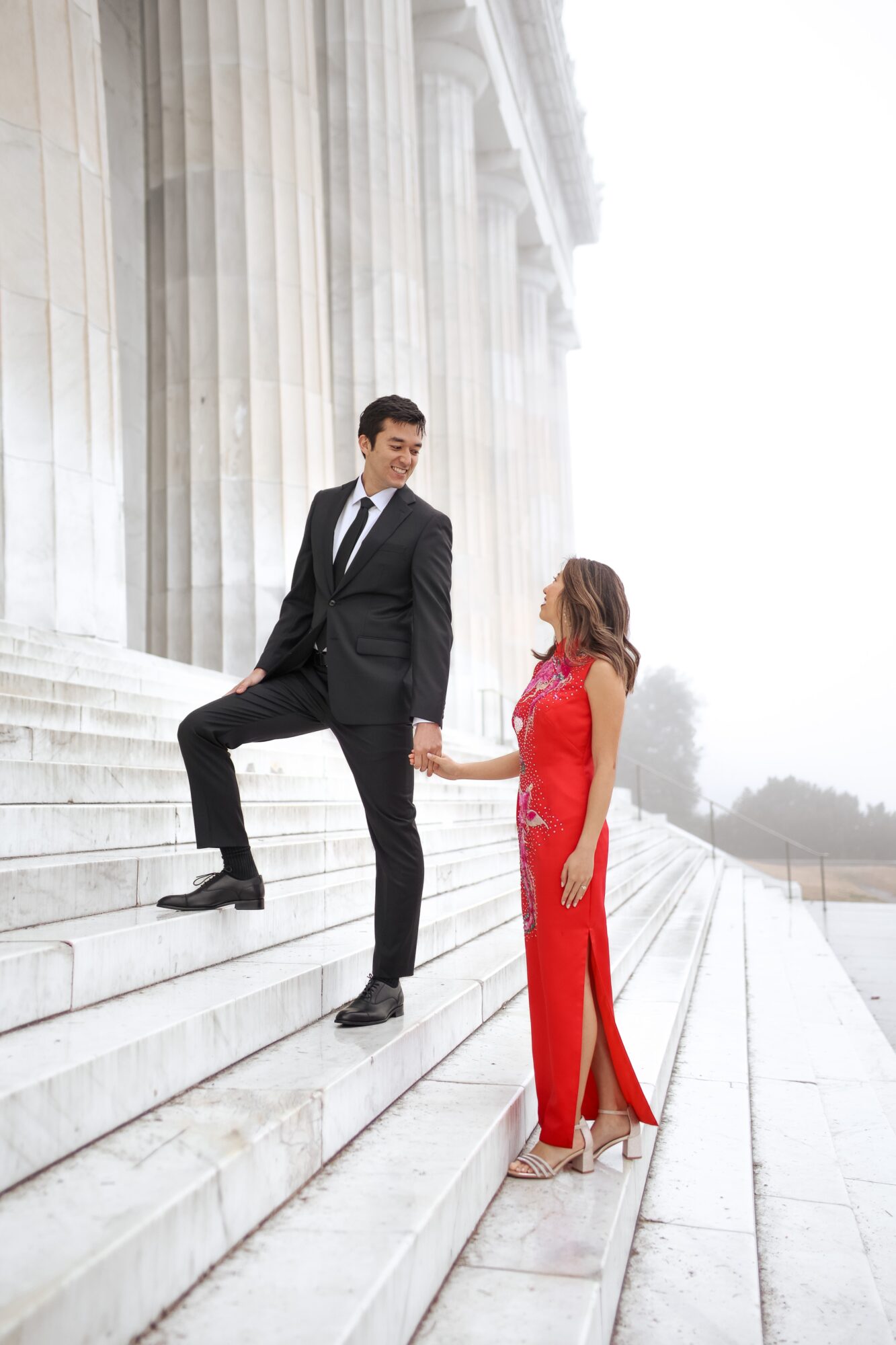 A man in a suit and a woman in a red dress stand on white steps outside a building with tall columns, holding hands.