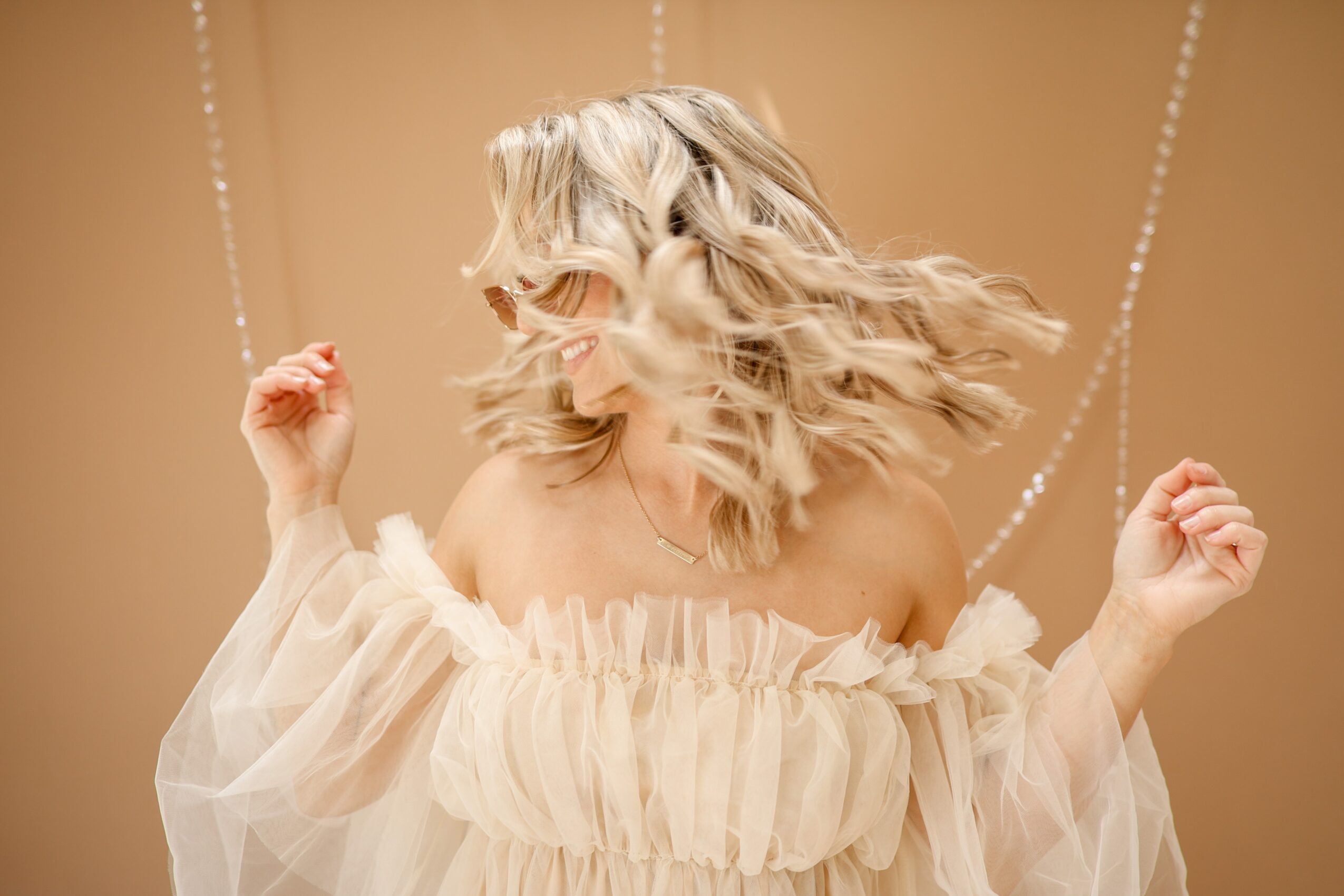 Woman with wavy blonde hair wearing an off-shoulder cream dress, smiling and swinging on a swing, background beige.