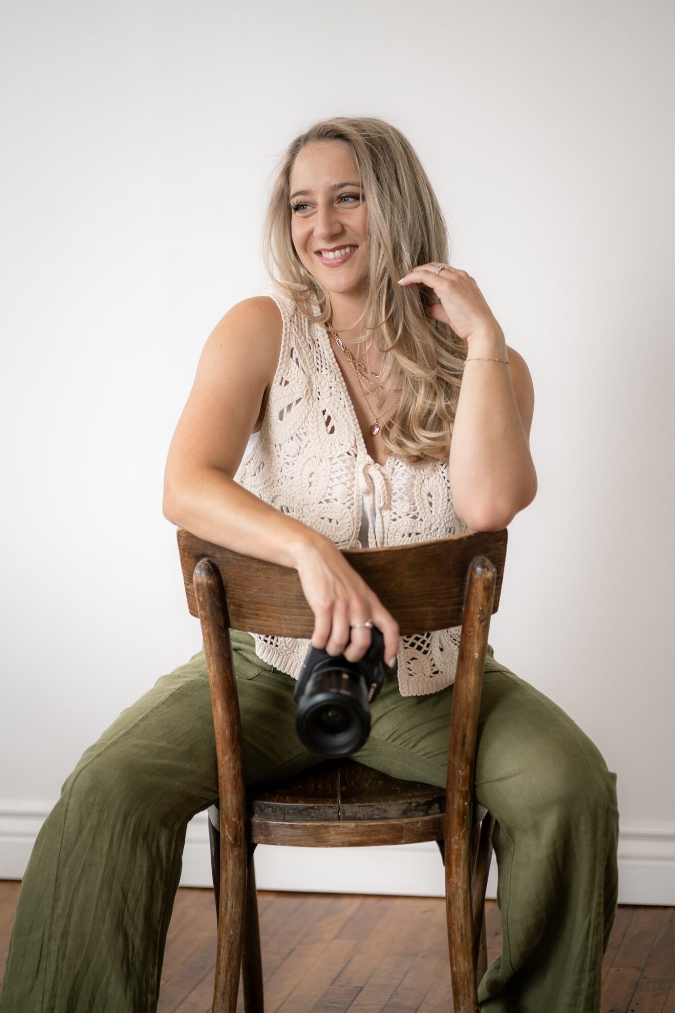 Woman sitting on a wooden chair, smiling, holding a camera, wearing a sleeveless top and green pants.