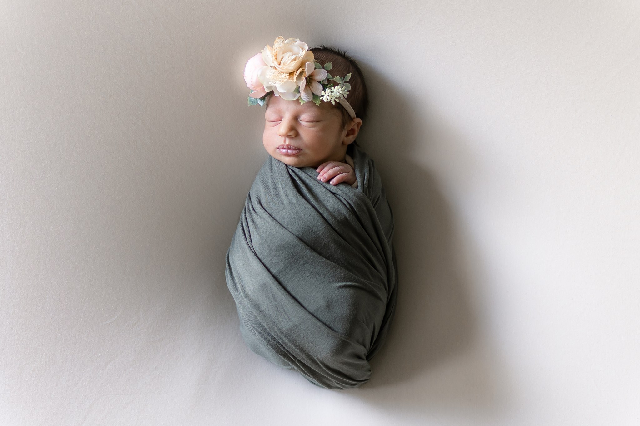 Newborn baby wrapped in gray cloth, wearing a floral headband, sleeping peacefully against a plain background.