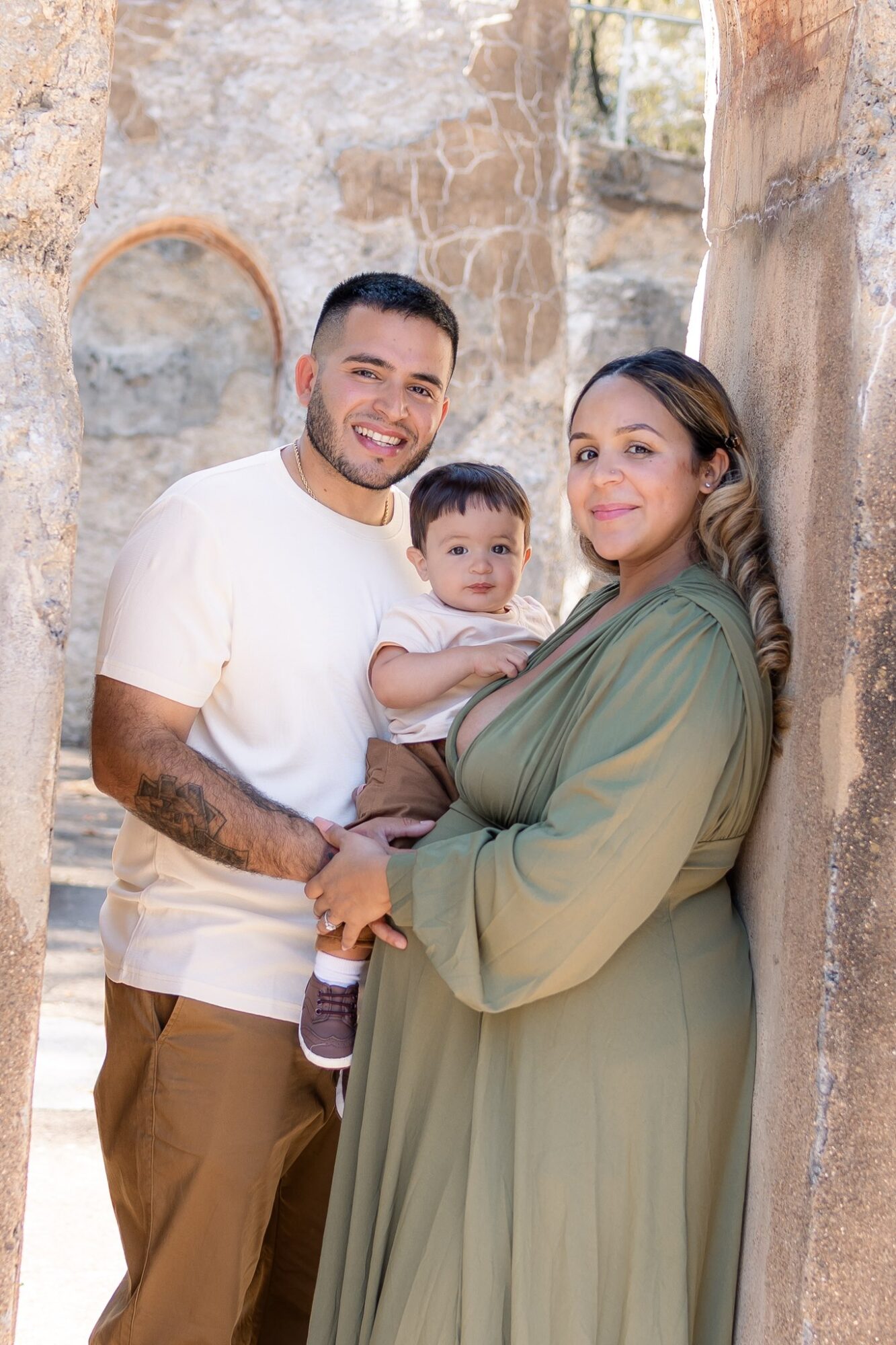 Family of three standing outdoors, smiling, with stone walls and arches in the background.