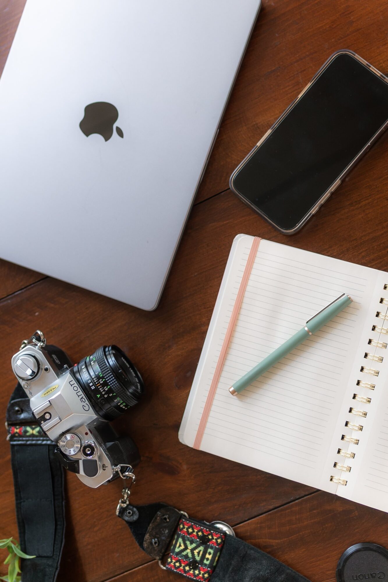 Laptop, smartphone, camera, and notebook with pen on wooden surface.