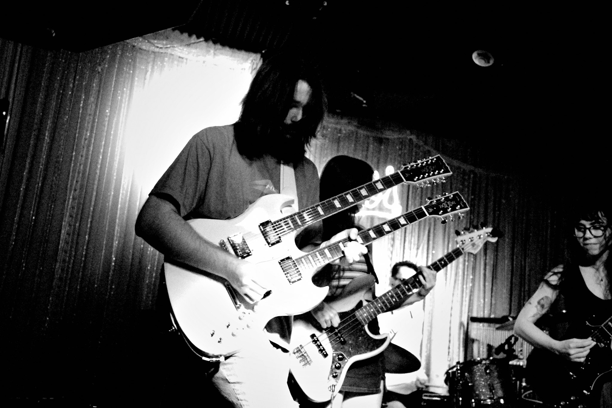 Three musicians playing electric guitars on stage with curtains in the background, in black and white.