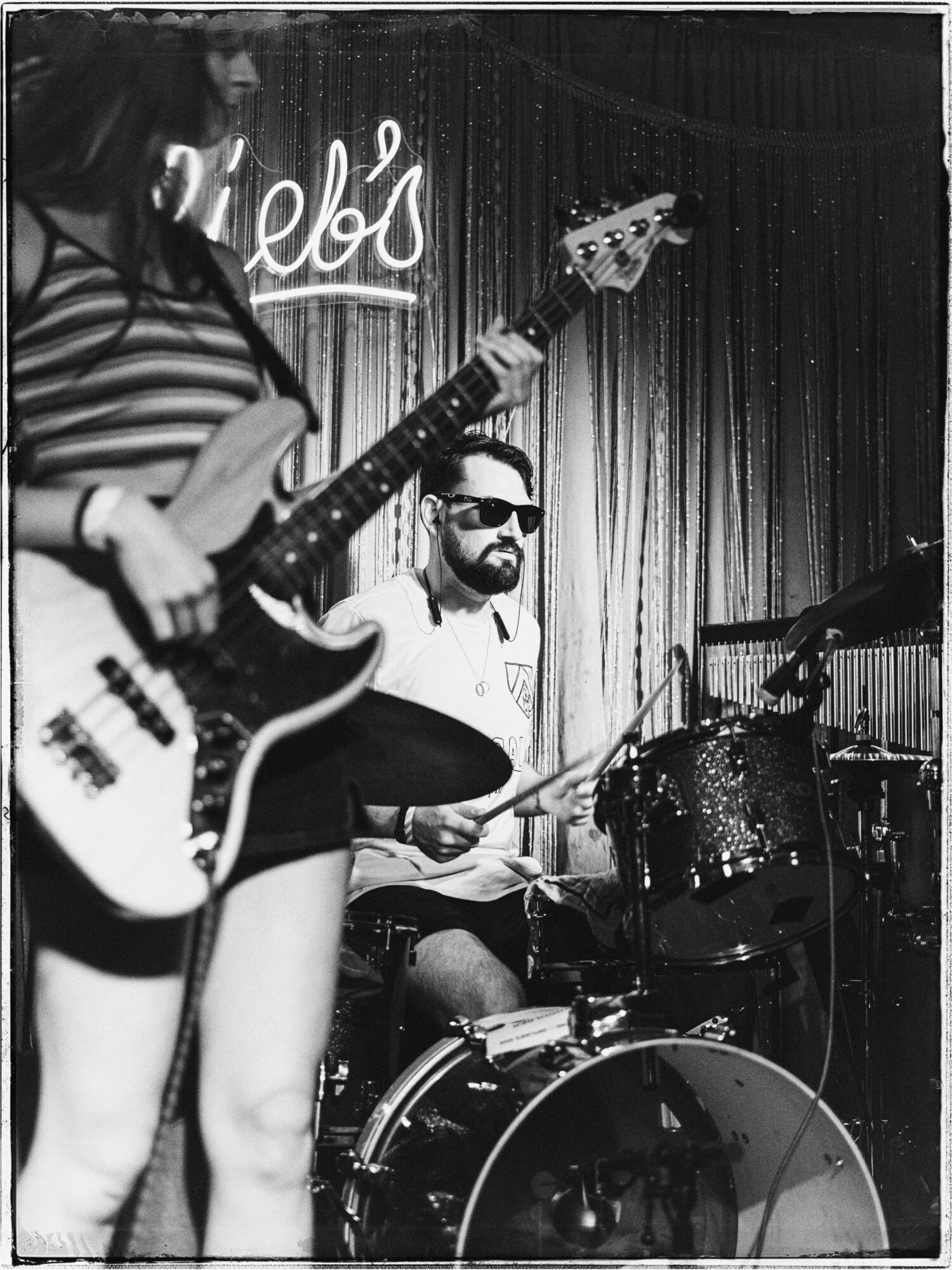 Two musicians perform on stage; one playing guitar, the other on drums, with a curtain backdrop and a neon sign.