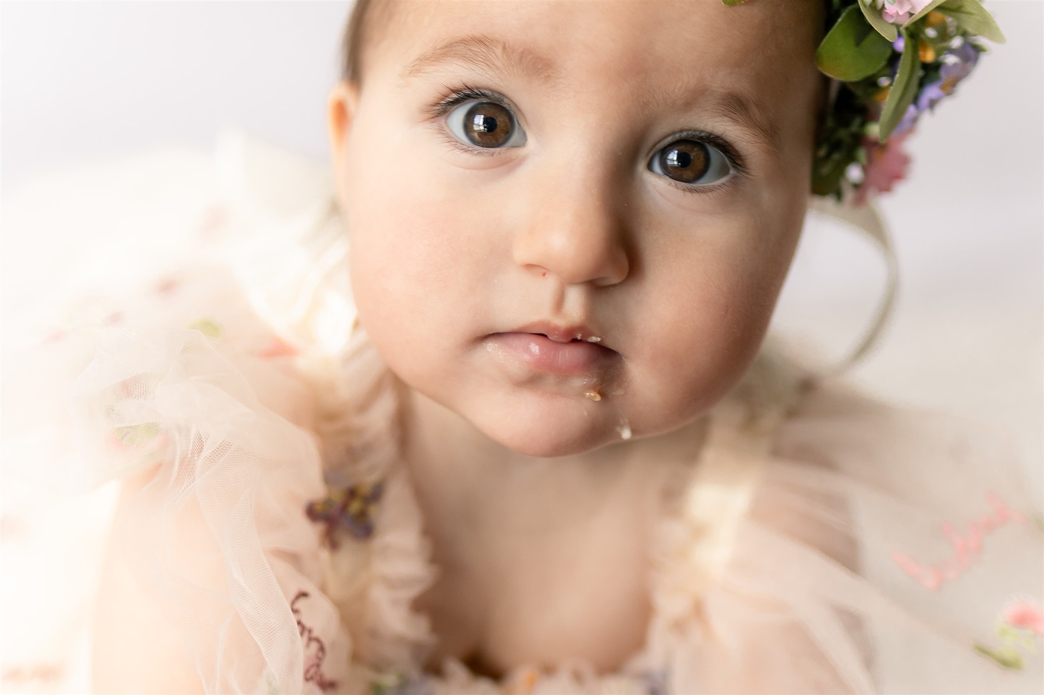 Close-up of a young child with big eyes, wearing a floral headband and a light-colored dress with ruffles.