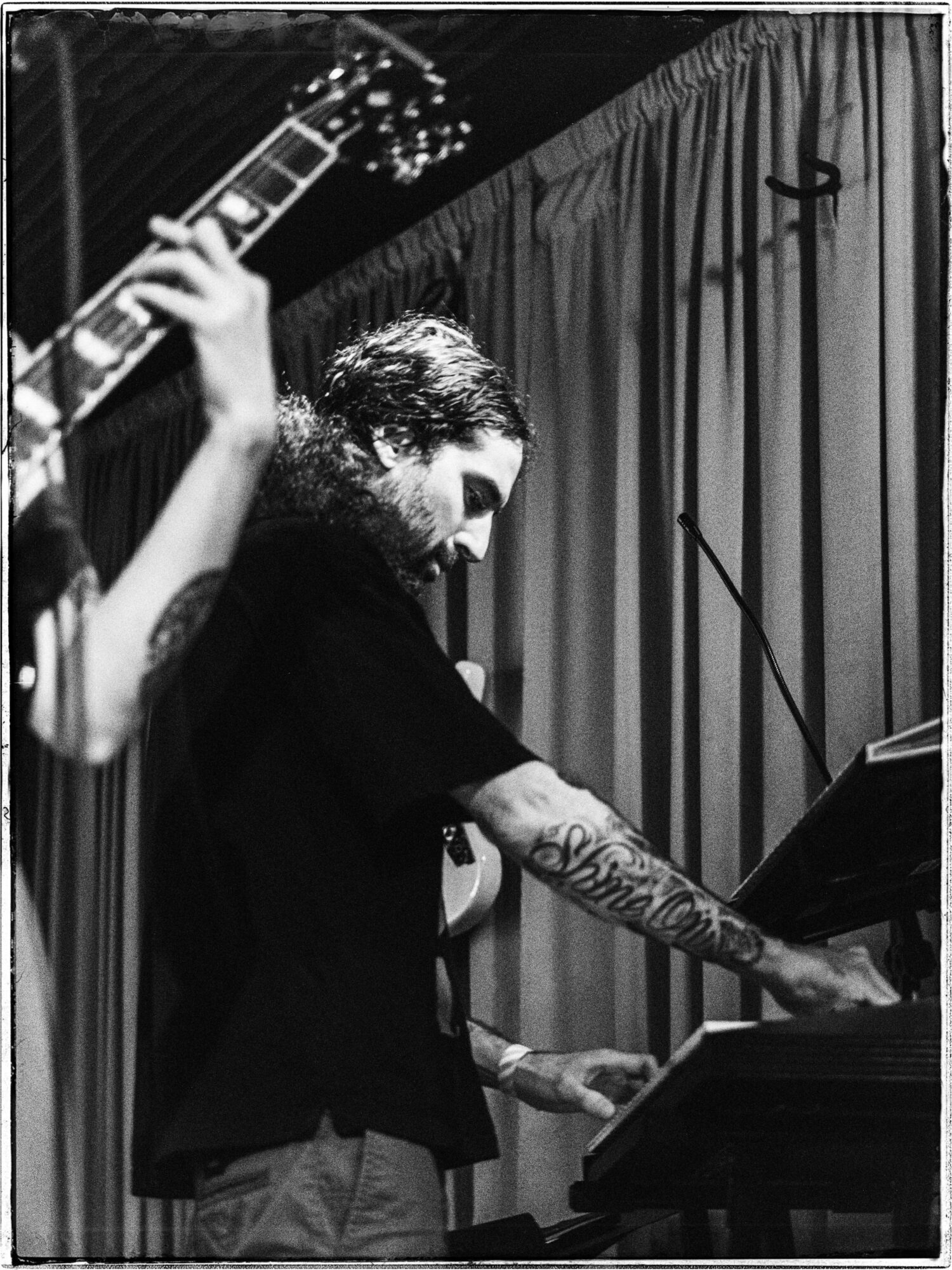 Person playing piano with a tattooed arm, curtain background, black and white photo.