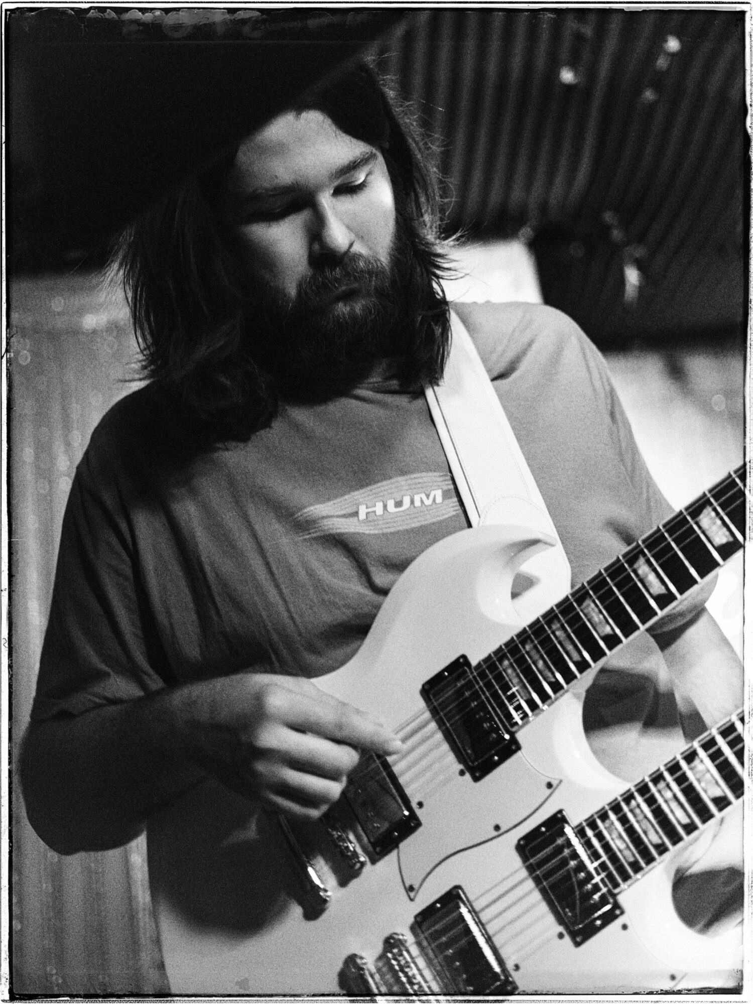 Person with long hair and beard playing a double-neck guitar, looking down, in a room with wooden walls.