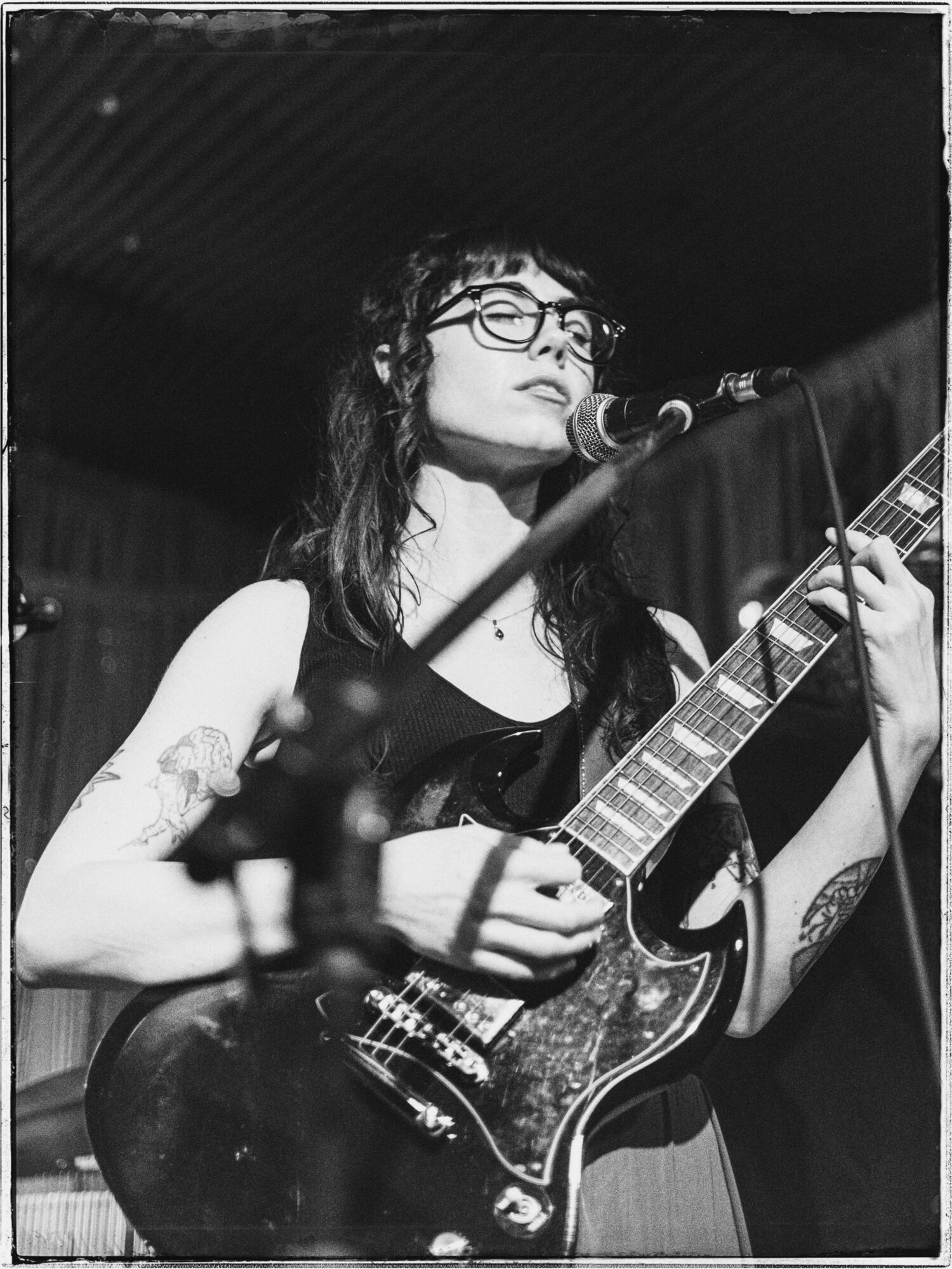 Woman with glasses playing an acoustic guitar on stage, singing into a microphone, in black and white.