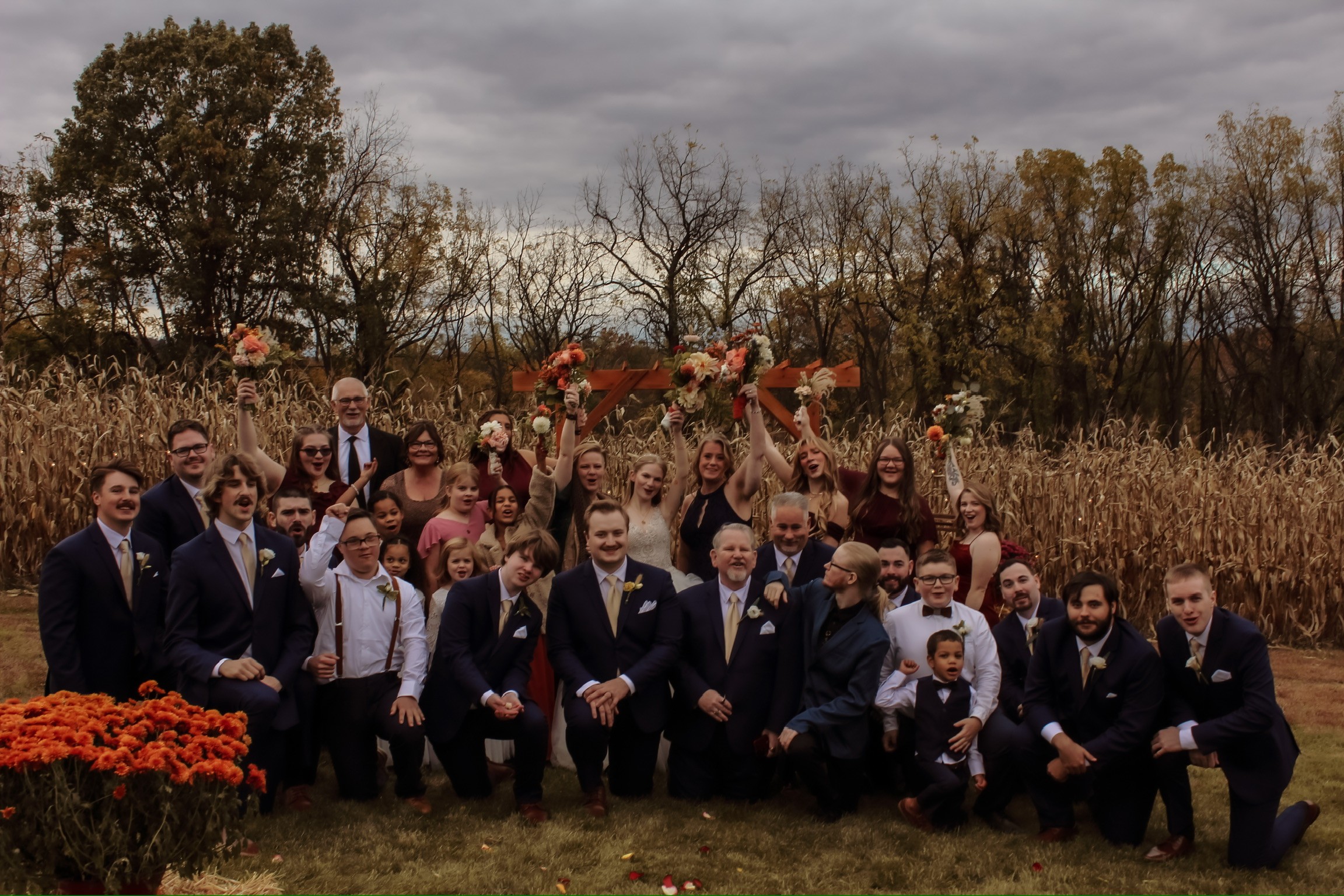 Group of people in formal attire outdoors with trees and cloudy sky in background.