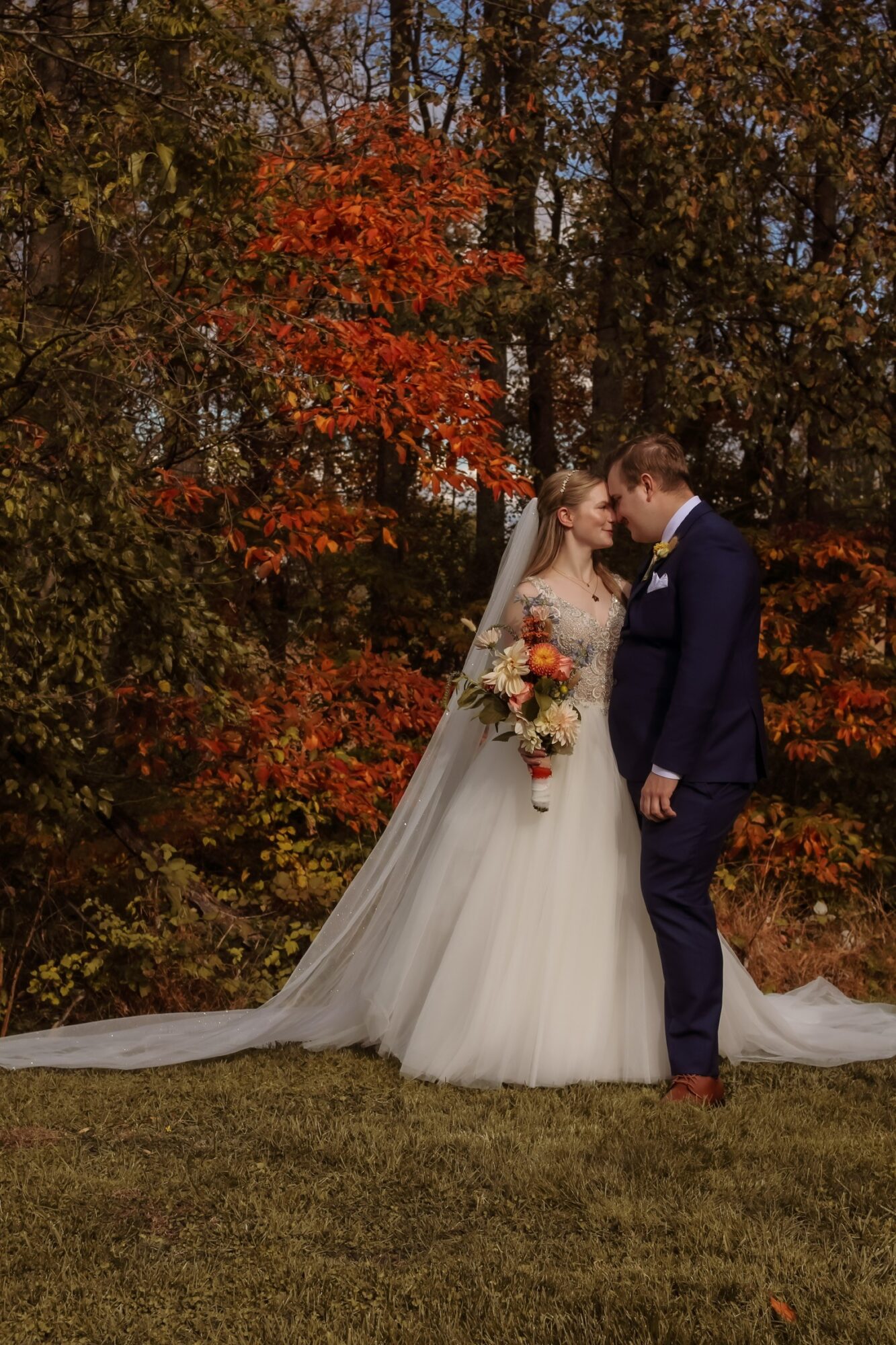 Bride and groom stand close, surrounded by autumn trees with colorful leaves, holding a bouquet of flowers.