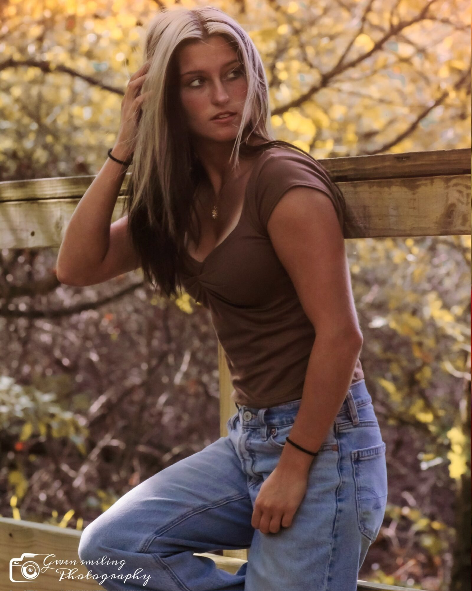 Young woman with long hair leaning on a wooden railing outdoors, surrounded by autumn trees with yellow leaves.
