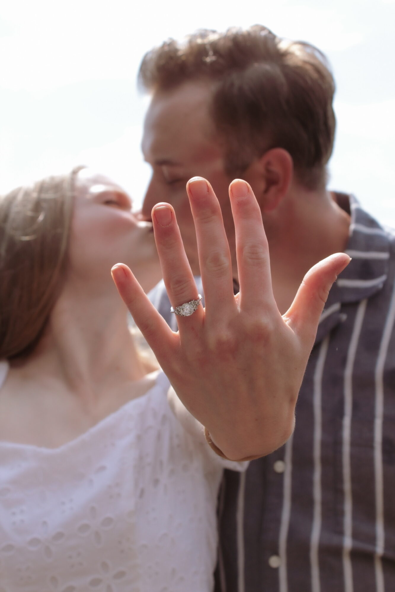 A woman shows her ringed hand in front of a man and woman kissing in the background.