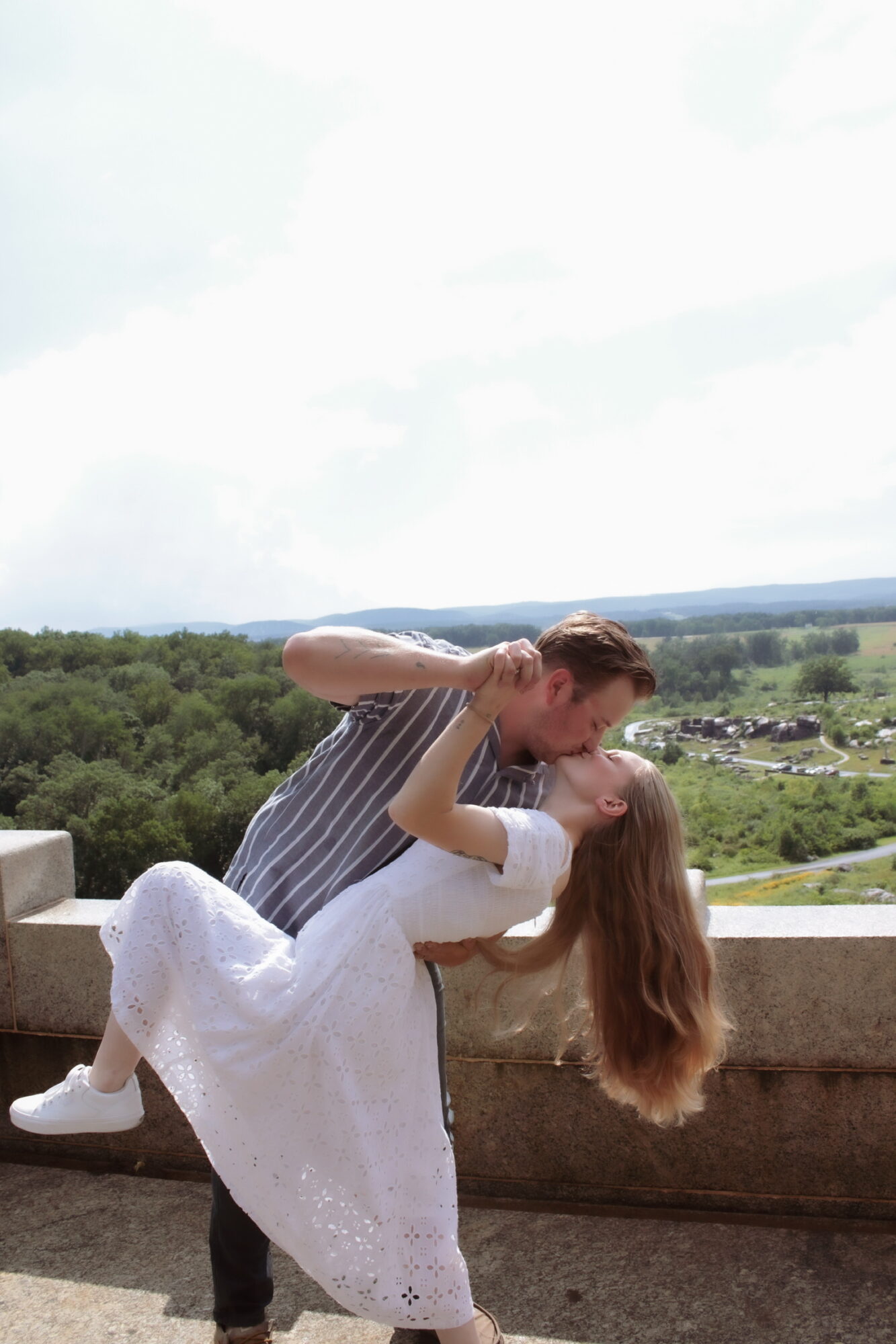 A couple kissing outdoors on a balcony with a landscape background, woman in white dress, man in striped shirt.
