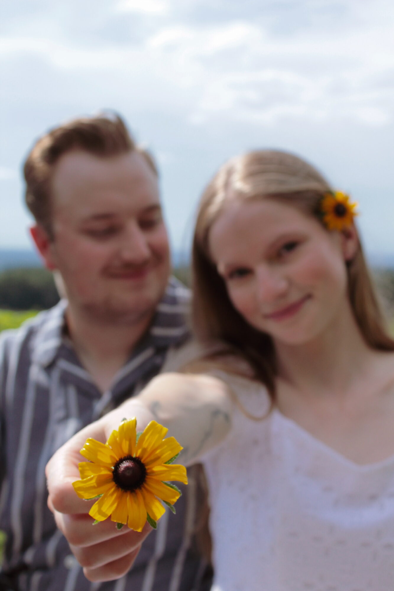 A young woman holds a yellow flower with a dark center, smiling at the camera, with a man in the background outdoors.