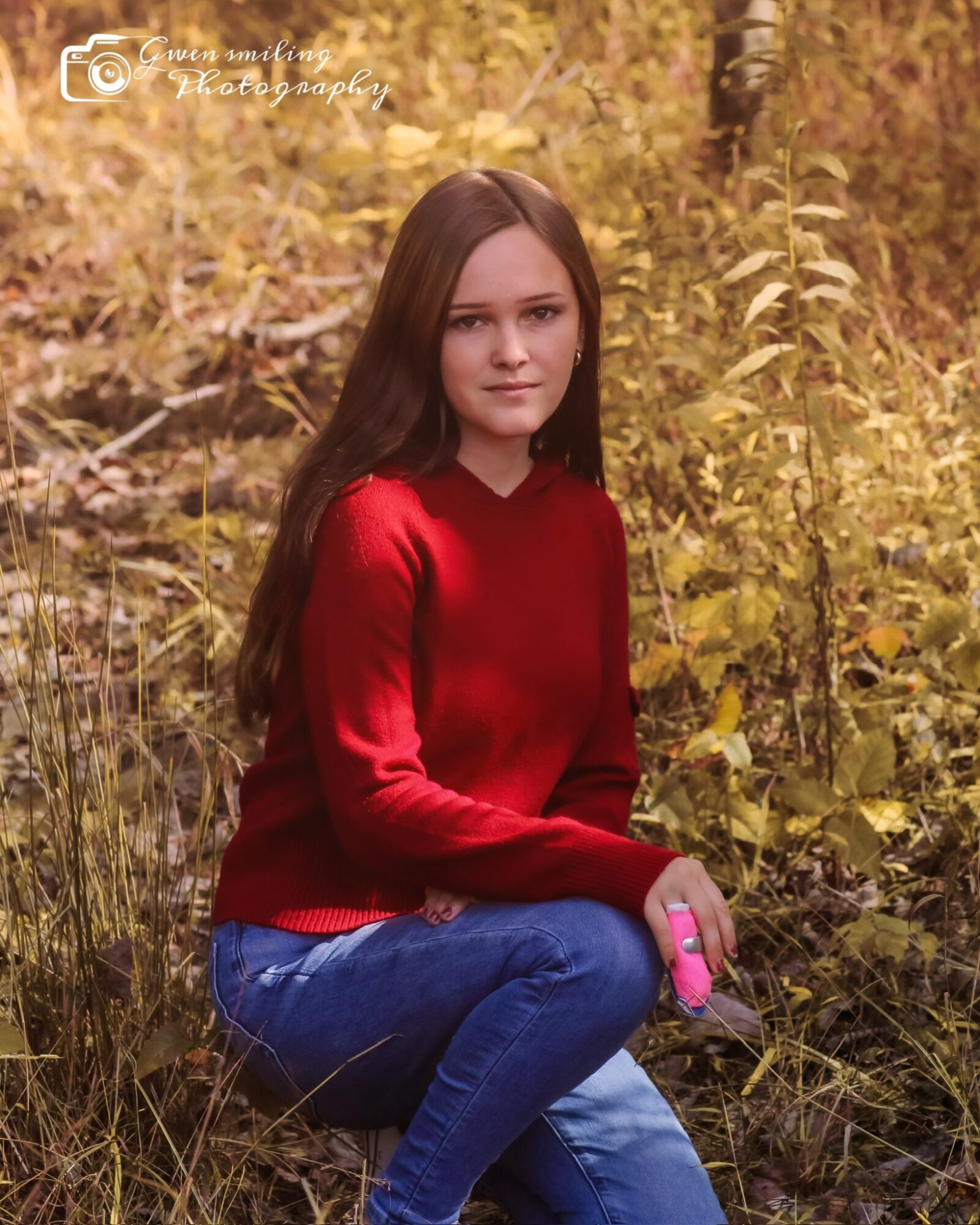 Young woman with long brown hair sitting outdoors among yellow foliage, wearing a red sweater and blue jeans, holding a pink object.