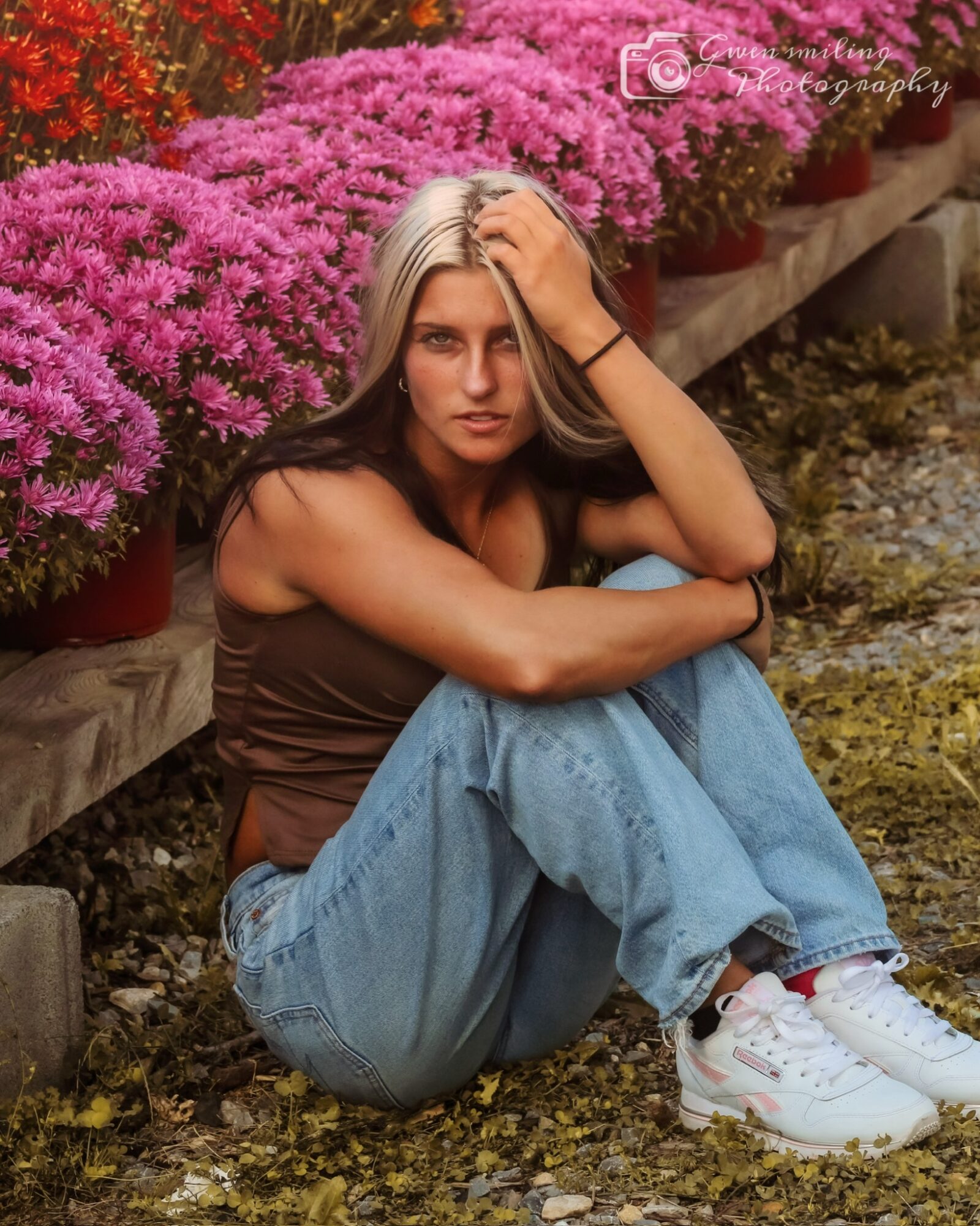 Young woman sitting outdoors near pink flowers, wearing a brown top, blue jeans, and white sneakers, with one hand on her head.