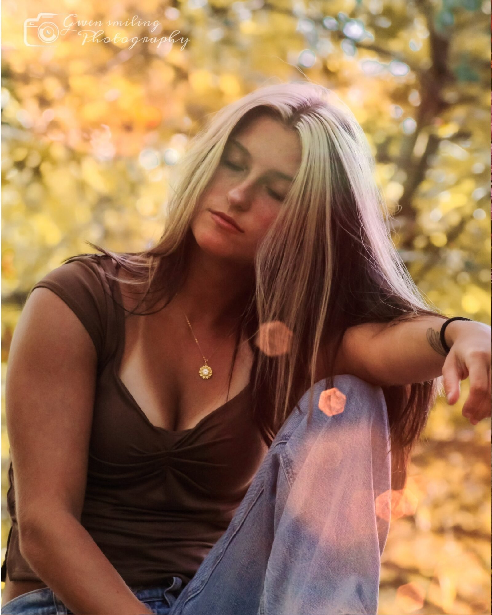Woman with long hair sitting outdoors with autumn leaves background, eyes closed, wearing a brown top and a necklace.