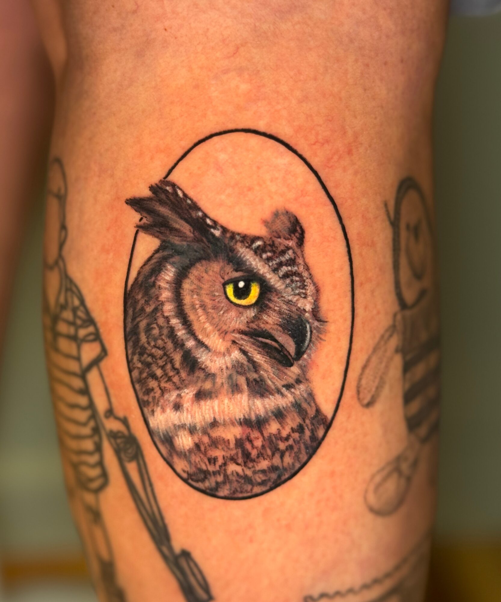 Tattoo of a cat's face with a bird's head inside an oval, with other tattoos partially visible around.